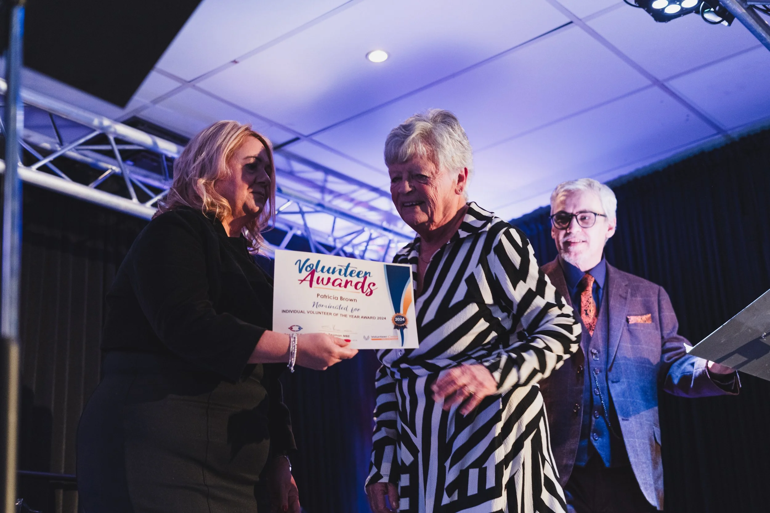 A woman receiving a Volunteer Awards certificate from a woman with blonde hair, wearing a black blazer and pants, on stage at an awards ceremony, with a man with glasses and gray hair standing in the background.
