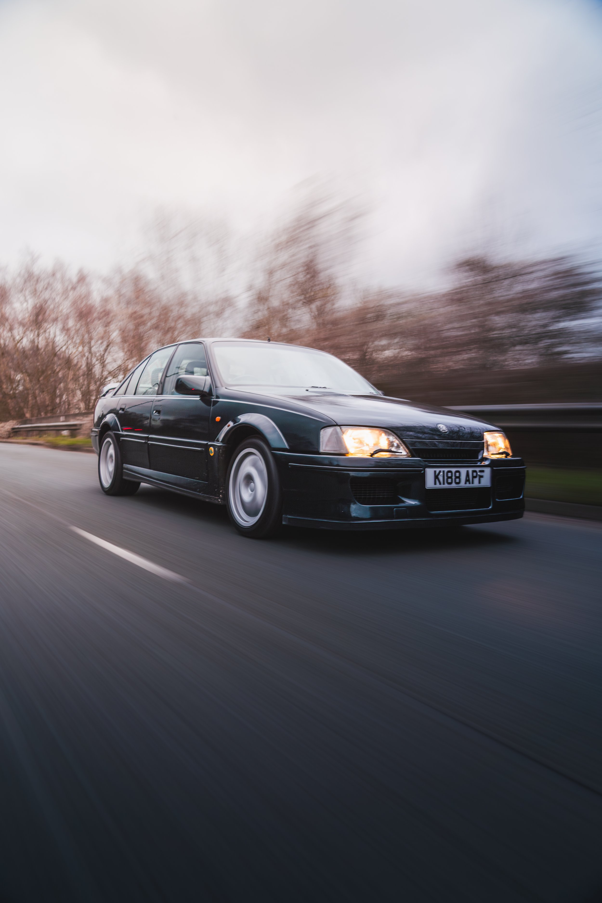 A dark-colored sedan car driving fast on a highway, with motion blur and cloudy sky in the background.