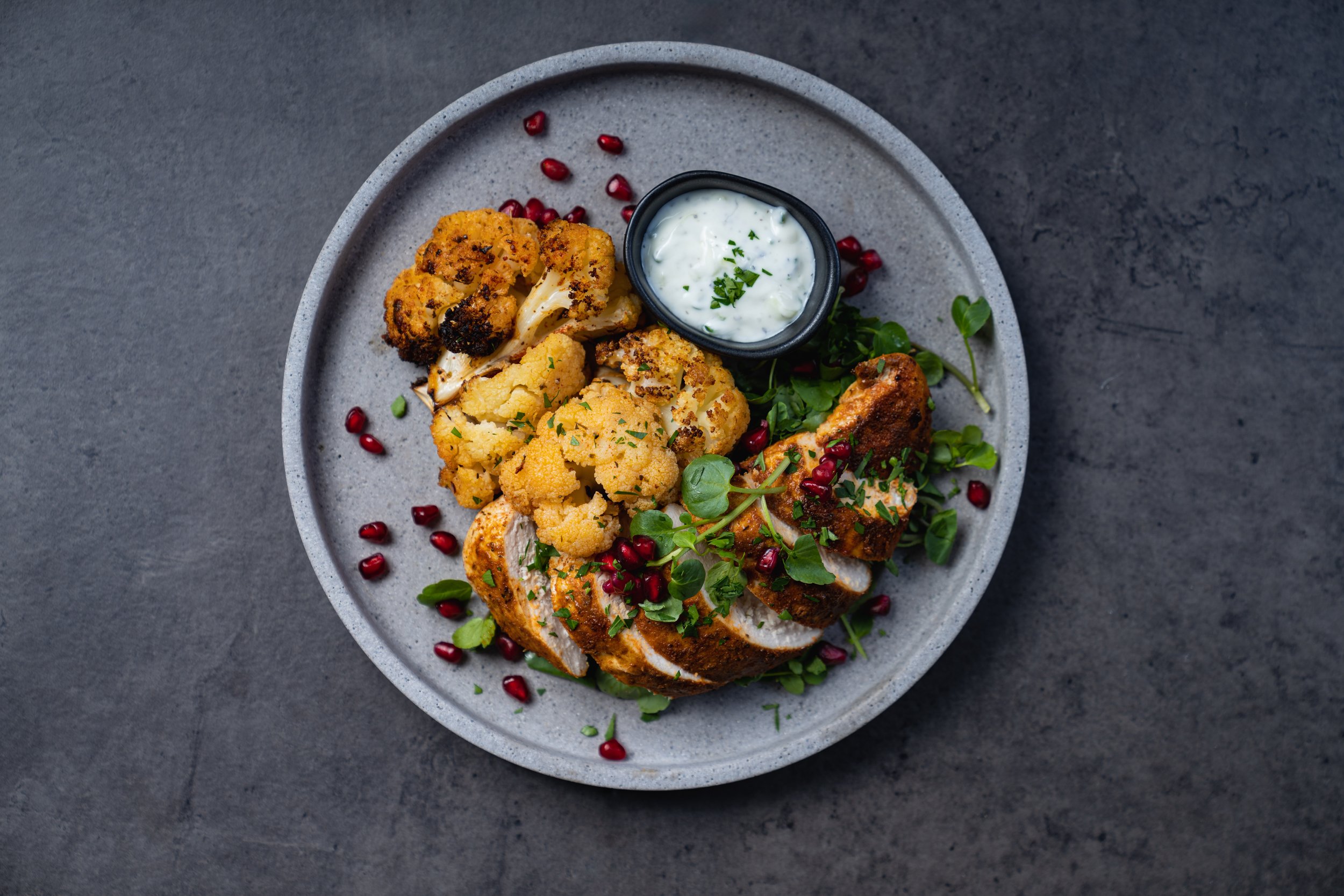 A plate of roasted cauliflower, grilled chicken breast topped with microgreens and pomegranate seeds, with a side of ranch dipping sauce.