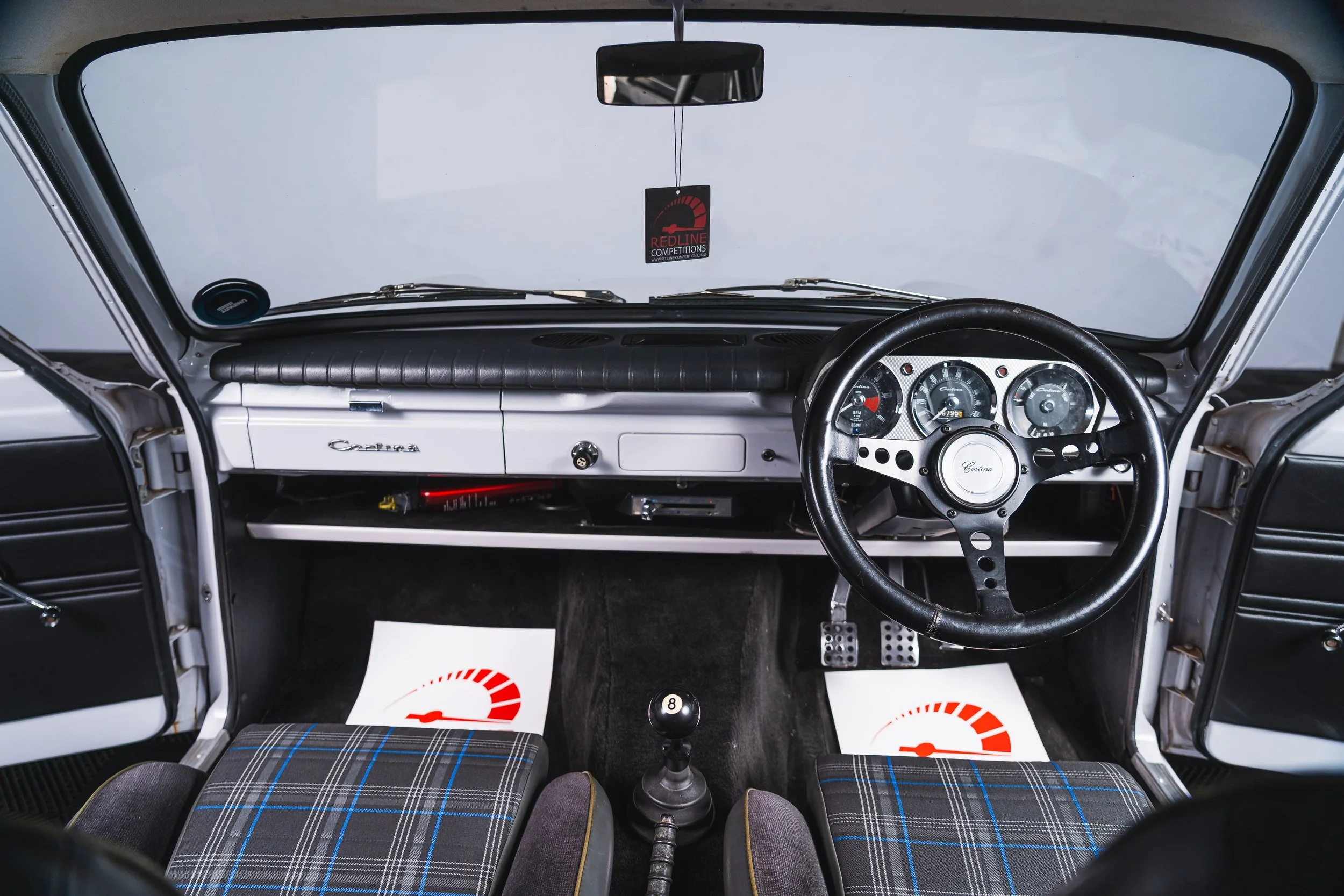 Interior view of a vintage car dashboard with a four-gauge cluster, steering wheel, manual gear shift knob marked with a number 8, plaid fabric seats, and a rearview mirror with a red and black shoe-shaped air freshener hanging.