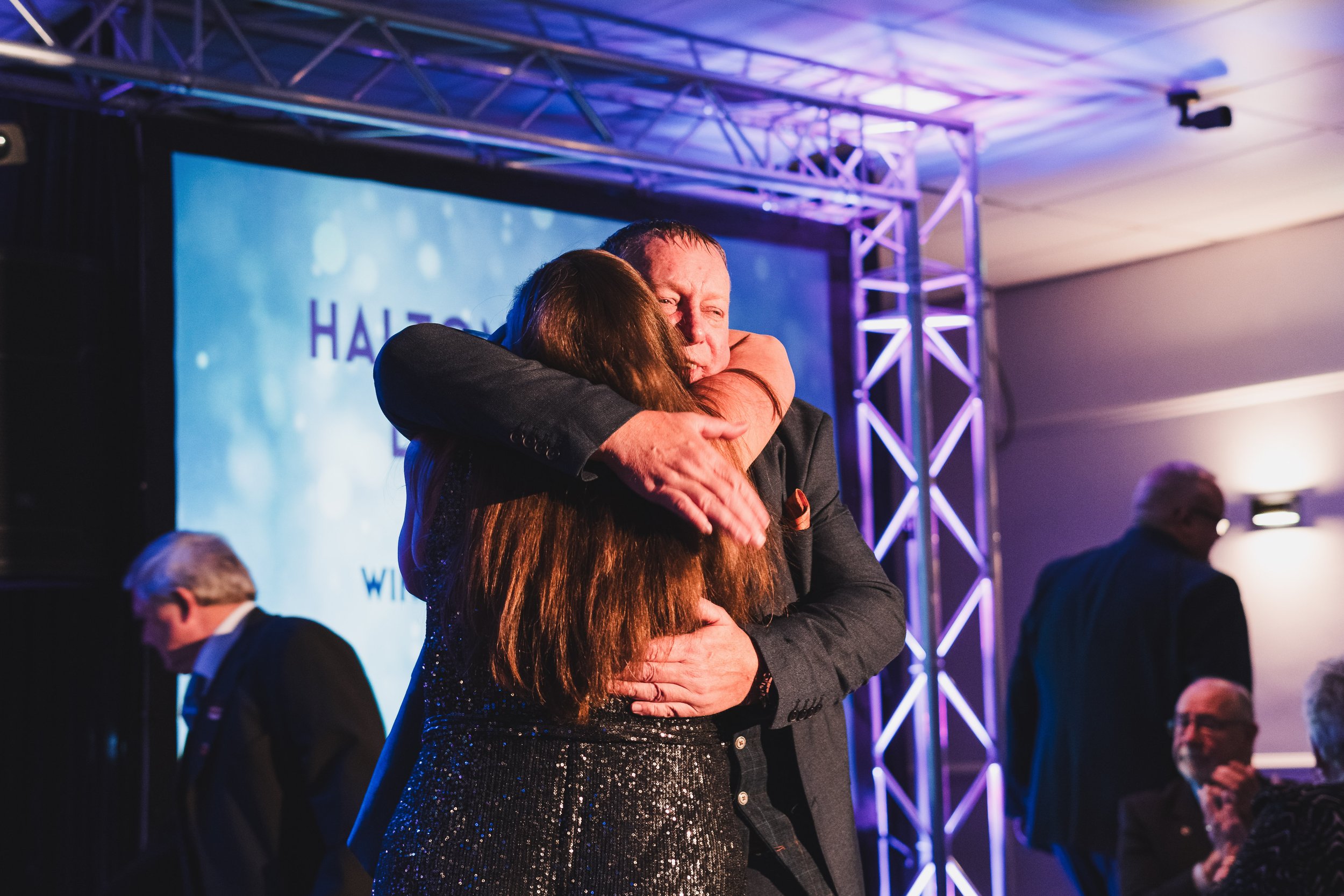 A man and woman hugging at an event with a stage and purple lighting in the background.