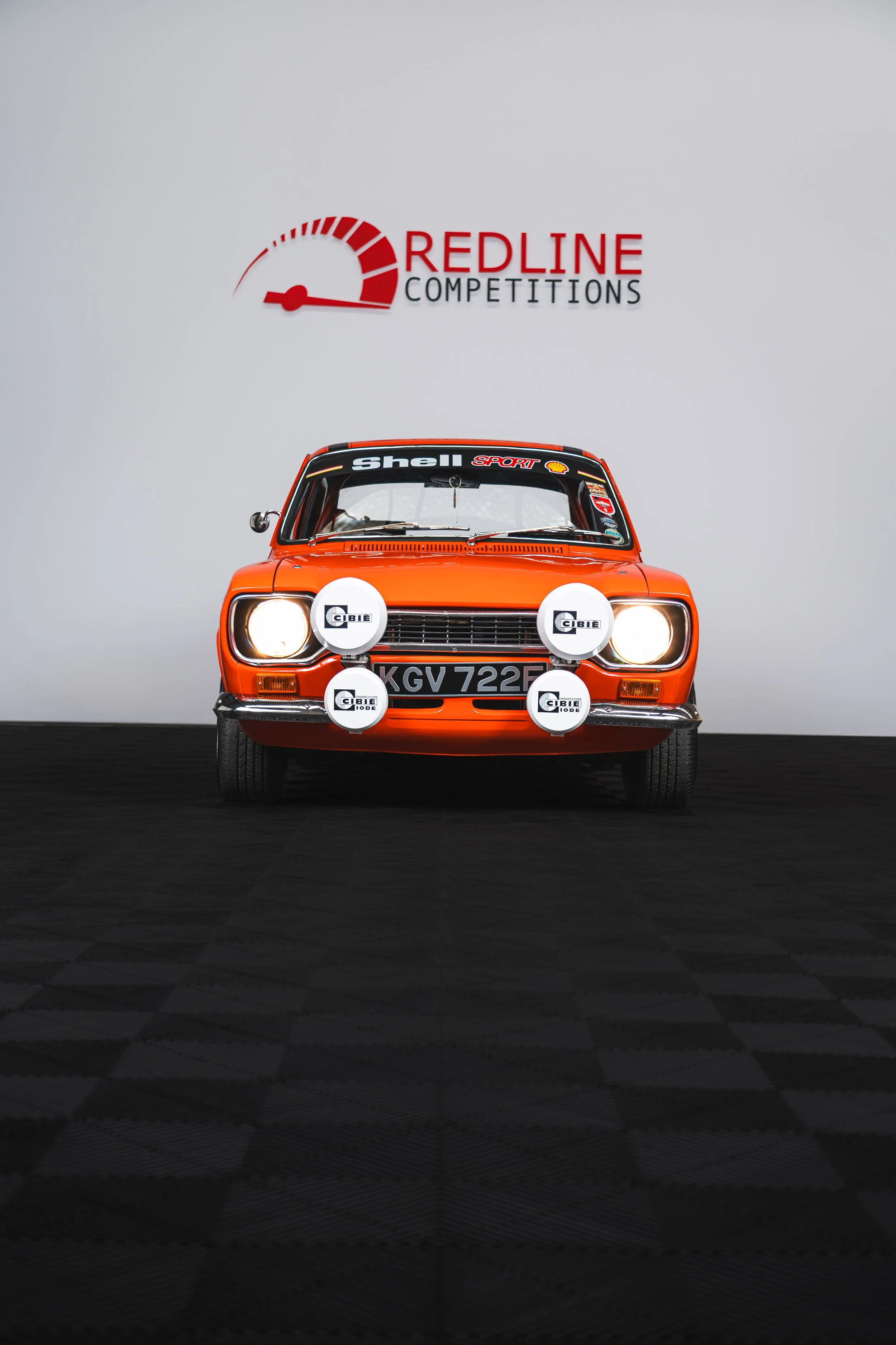Front view of an orange classic race car with four additional headlights, parked in front of a white wall with a "Redline Competitions" logo.