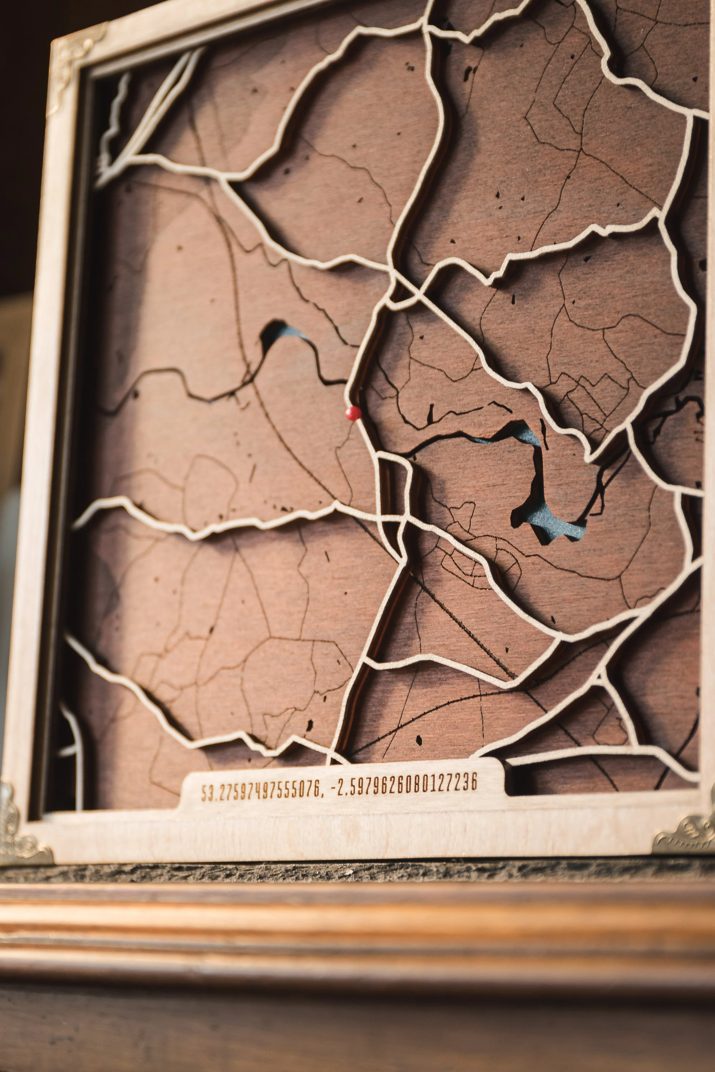 Close-up of a wooden framed map with irregular black outlines of regions or countries, a small red dot, and geographical coordinates printed on a metal plate at the bottom.