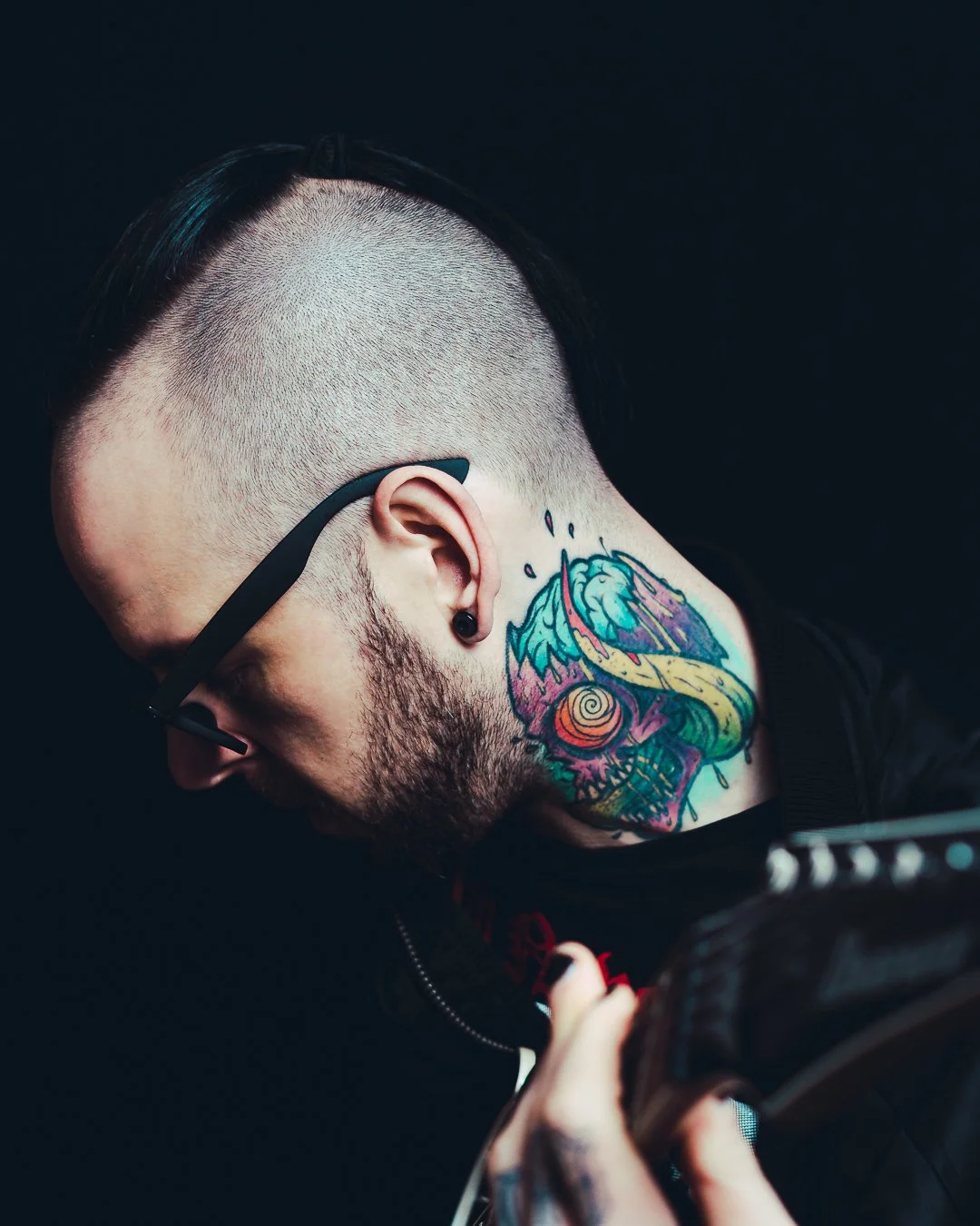 A man with a tattoo on his neck, wearing glasses, with a shaved side of his head, playing an electric guitar.