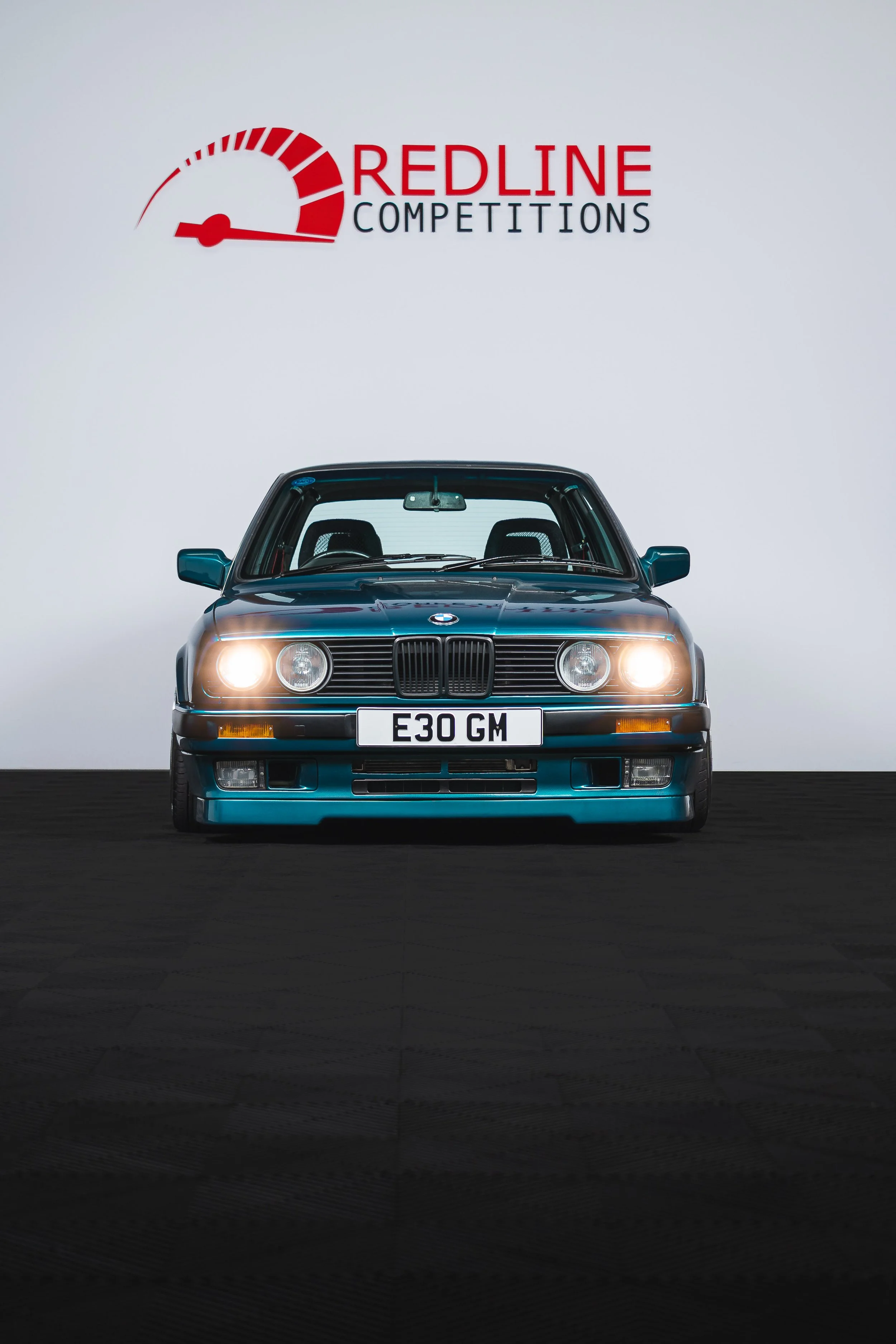 A blue classic BMW E30 car with headlights on, displayed at a Redline Competitions event against a plain background with the company's logo.
