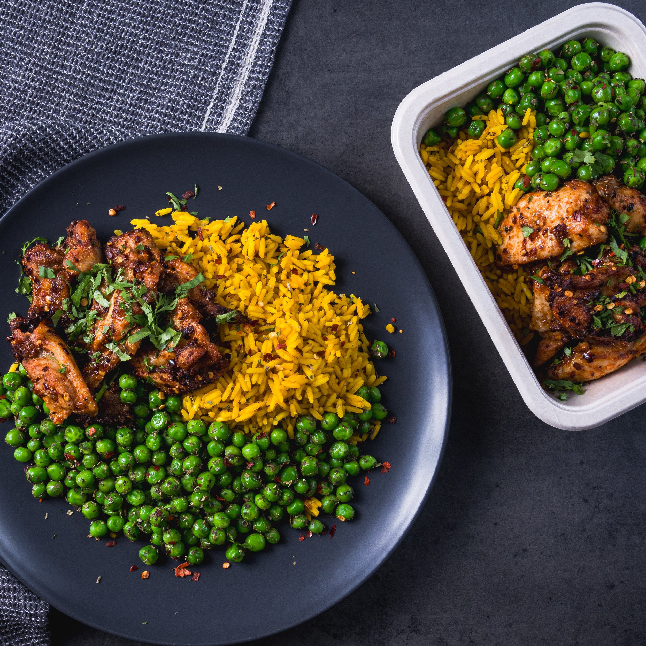Plate and container of chicken with cooked green peas and yellow rice, garnished with herbs, on a dark surface.