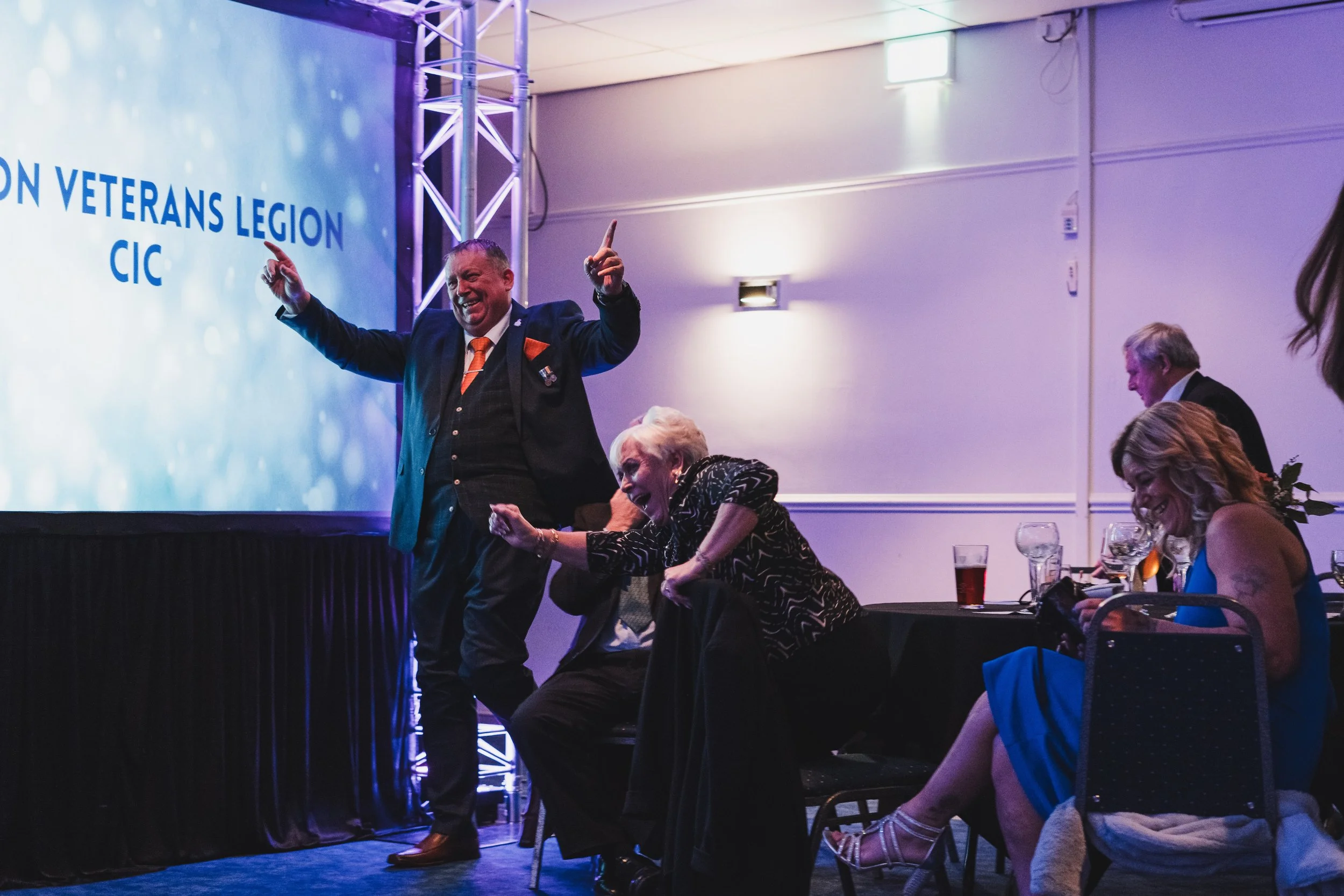 A man in a suit with a pin on his lapel is standing on stage, smiling and raising his arms in celebration. Two women sitting at a table are laughing and clapping, with drinks in front of them. The background shows a large screen with the words 'VETER