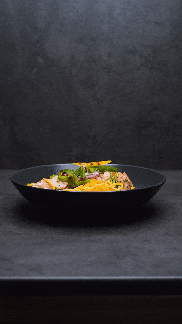 A black plate of Asian-style noodles with sliced chicken, green onions, red onions, and a yellow garnish, placed on a dark surface with a dark background.