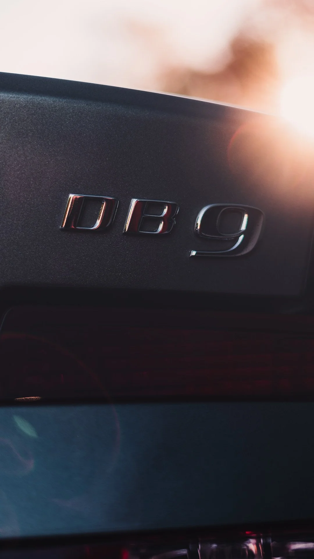 Close-up of a black car trunk with chrome emblem saying 'D B 9', sunlight in the background.