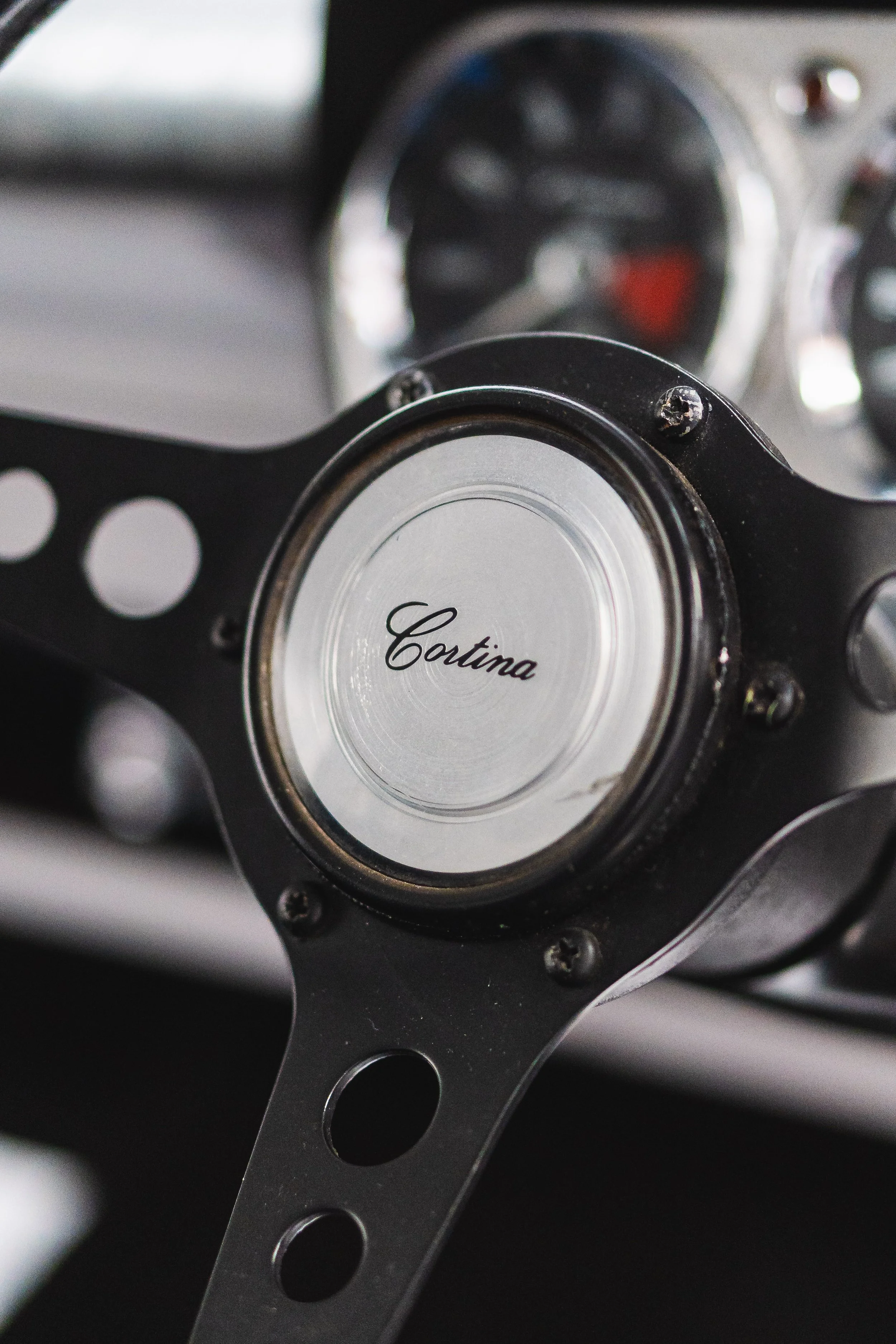 Close-up of a vintage car steering wheel with a metal center labeled 'Coutina', with dashboard gauges visible in the background.