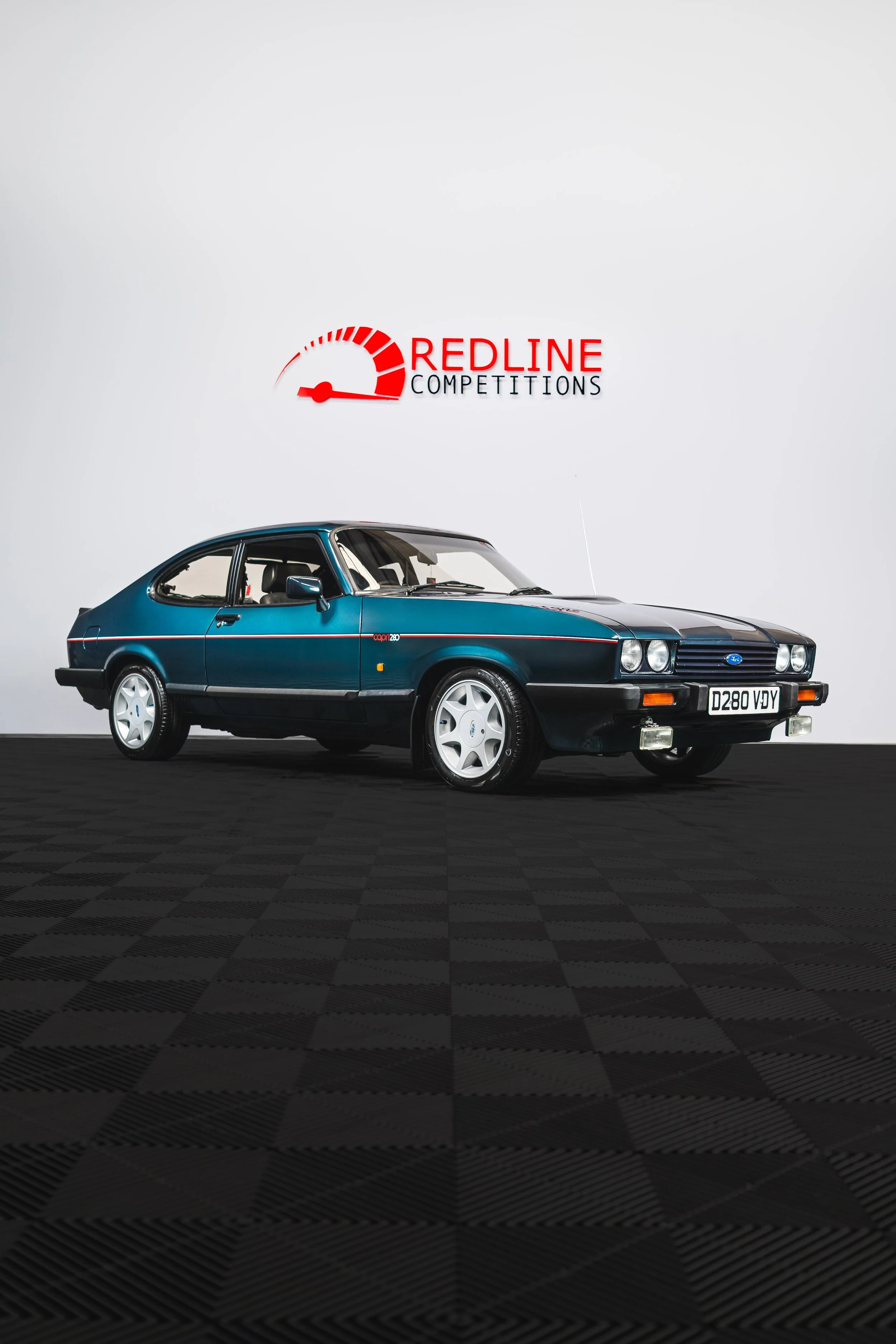 A classic blue Ford Capri car displayed in front of a white wall with a 'Redline Competitions' logo and a speedometer graphic.