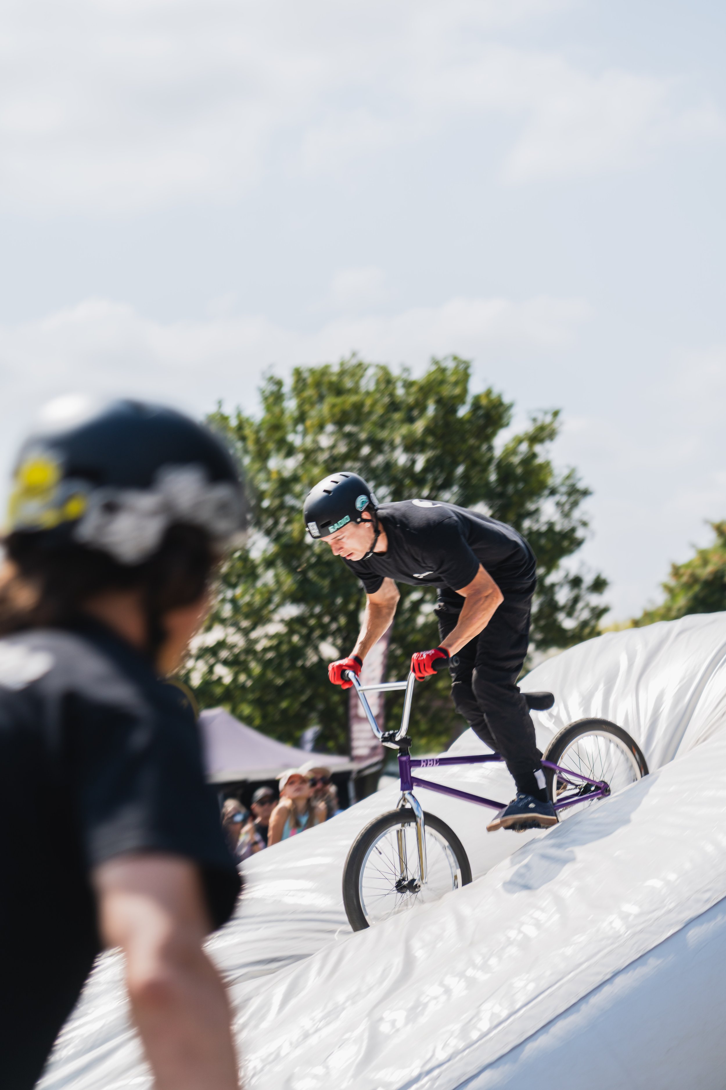 A person riding a BMX bike on a snowy ramp during a snowboarding event, with spectators watching in the background.
