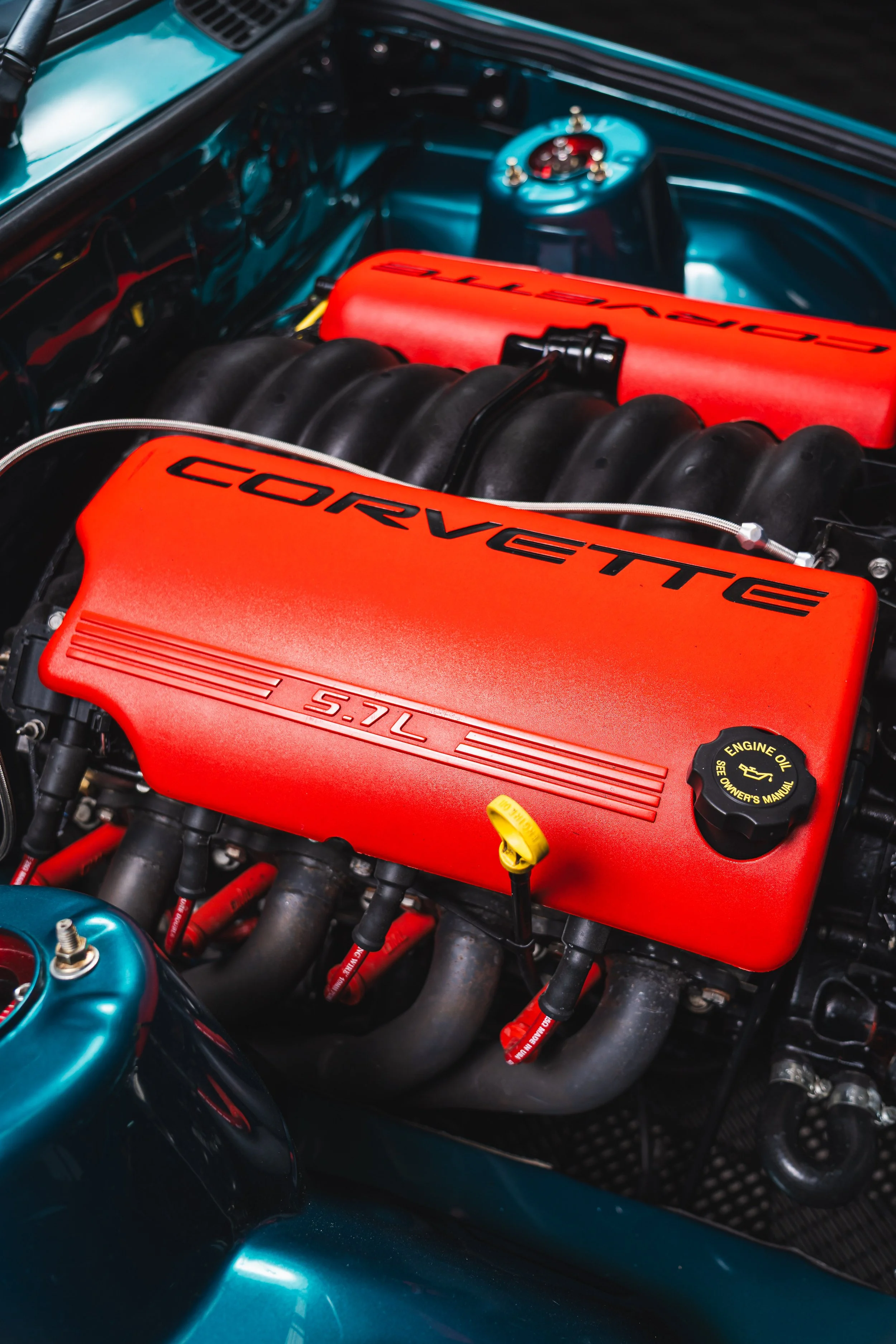 Close-up view of a Chevrolet Corvette engine with a bright red valve cover labeled 'CORVETTE 5.7L' and a yellow oil cap, surrounded by black hoses and metallic engine components.