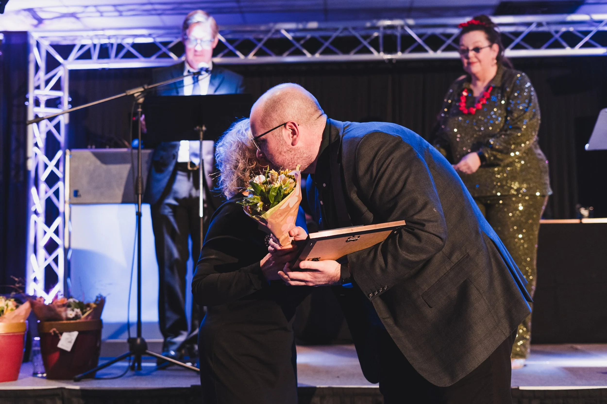 A man giving a woman a flower bouquet and a framed certificate or award on stage, with a band playing in the background.