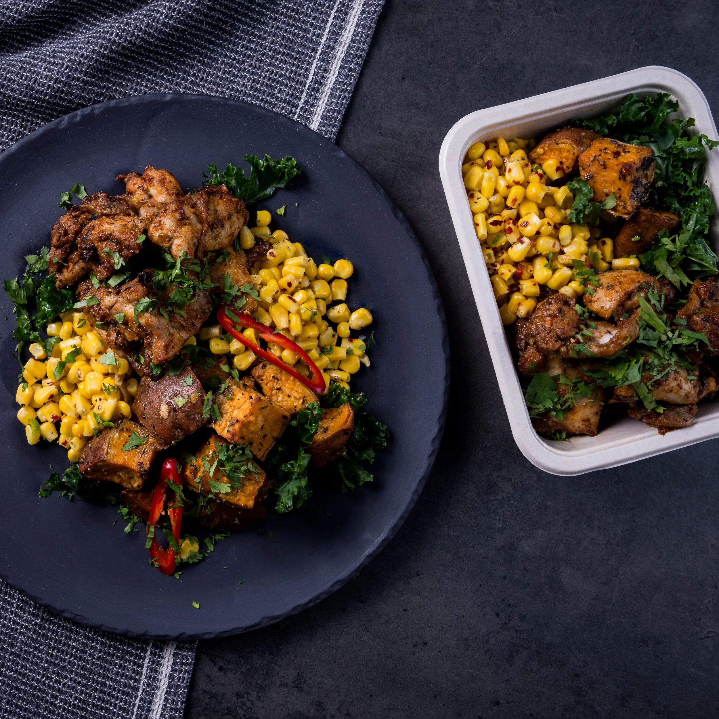 A black plate with grilled chicken, roasted butternut squash, sautéed kale, corn, and sliced red chili peppers; a white container filled with similar food items on a dark surface.