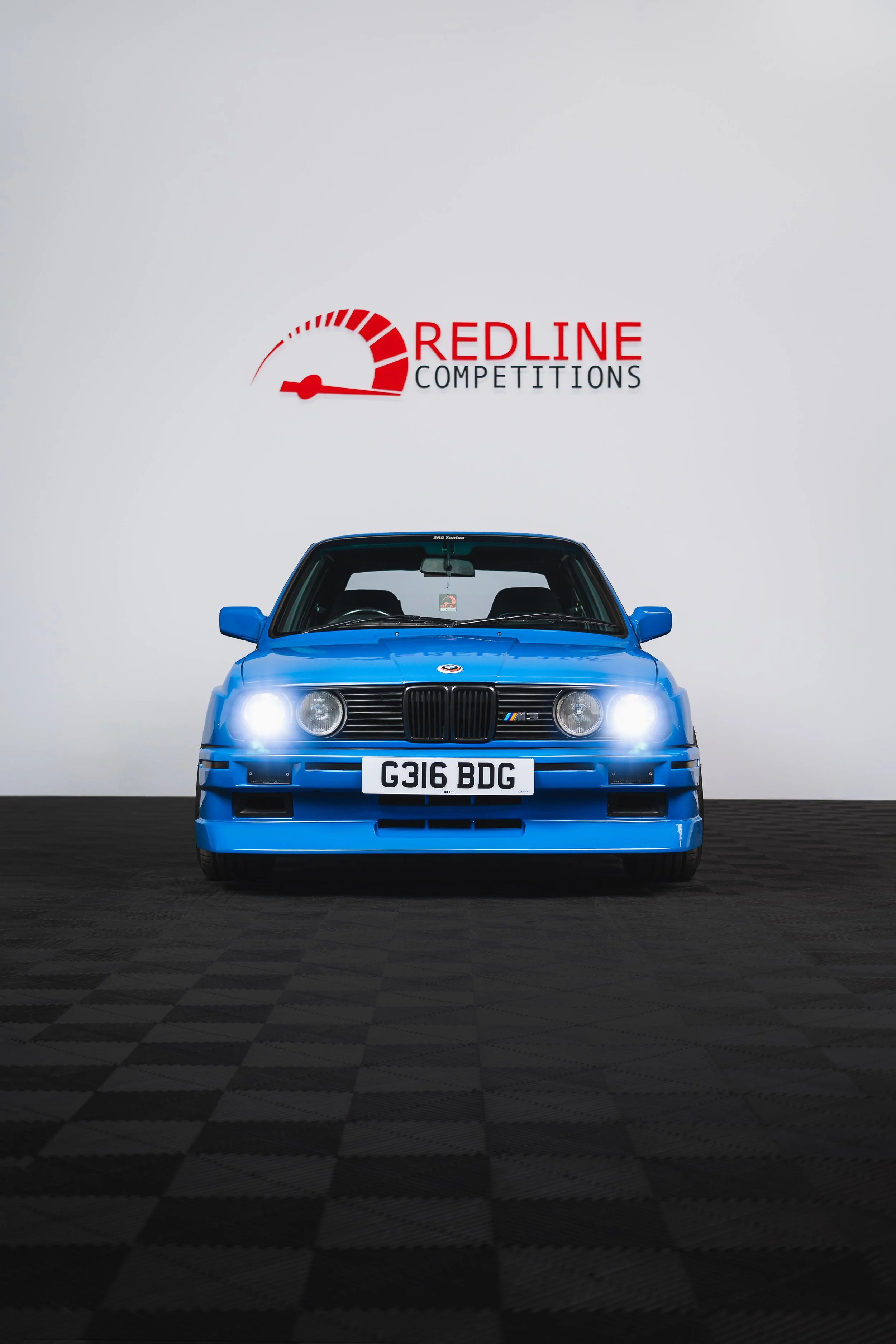 Blue BMW E30 M3 parked indoors with headlights on, in front of a white wall with a red 'Redline Competitions' logo and a speedometer graphic.