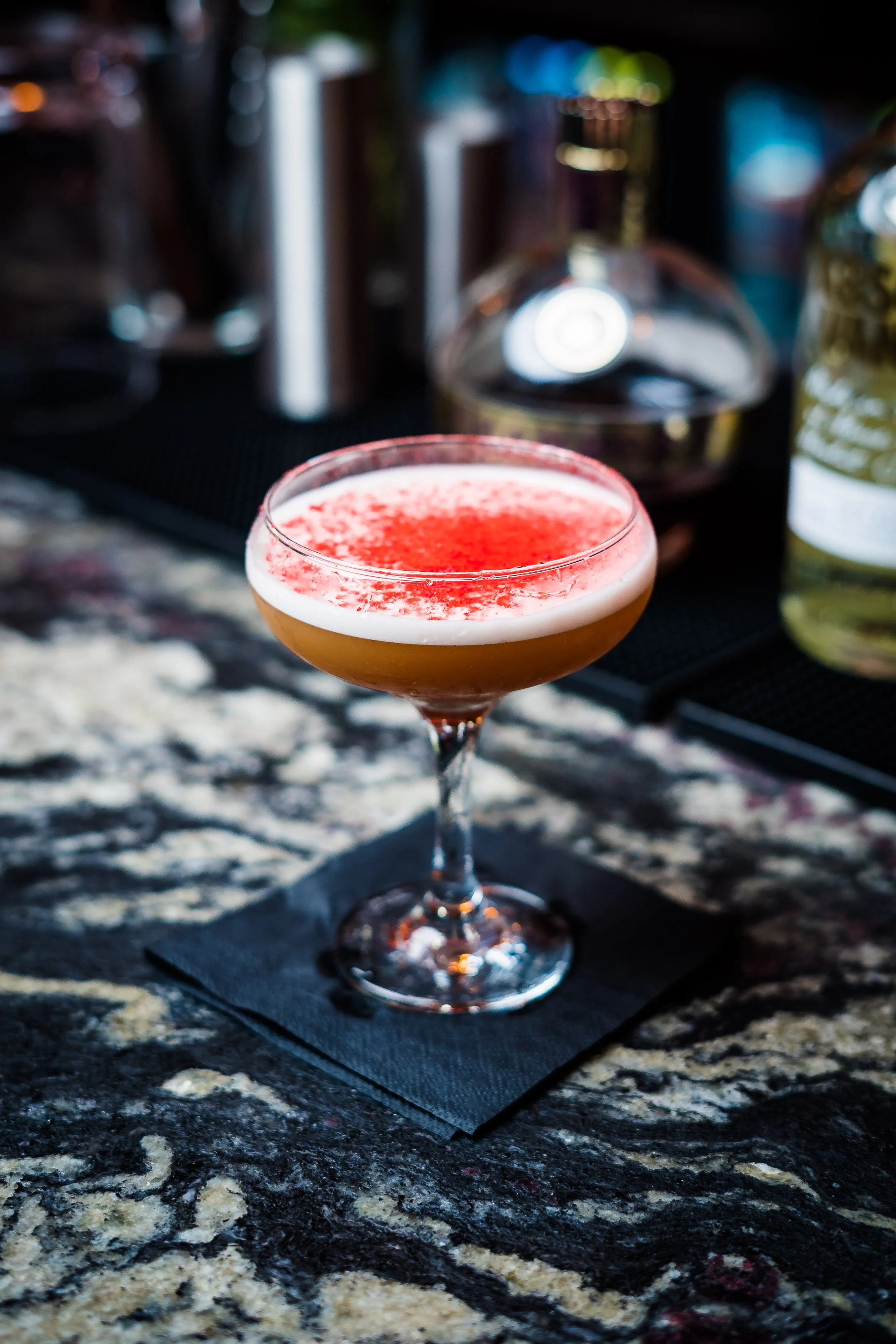 A cocktail glass with a layered drink topped with foam and red garnish, placed on a black napkin on a dark marble countertop, with bar tools and bottles in the background.