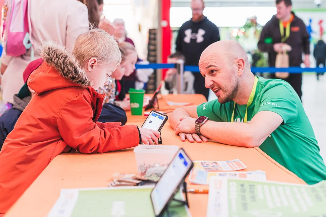 A man in a green shirt talking to a young boy in an orange jacket at a table with flyers and smartphones, with other children and adults in the background.