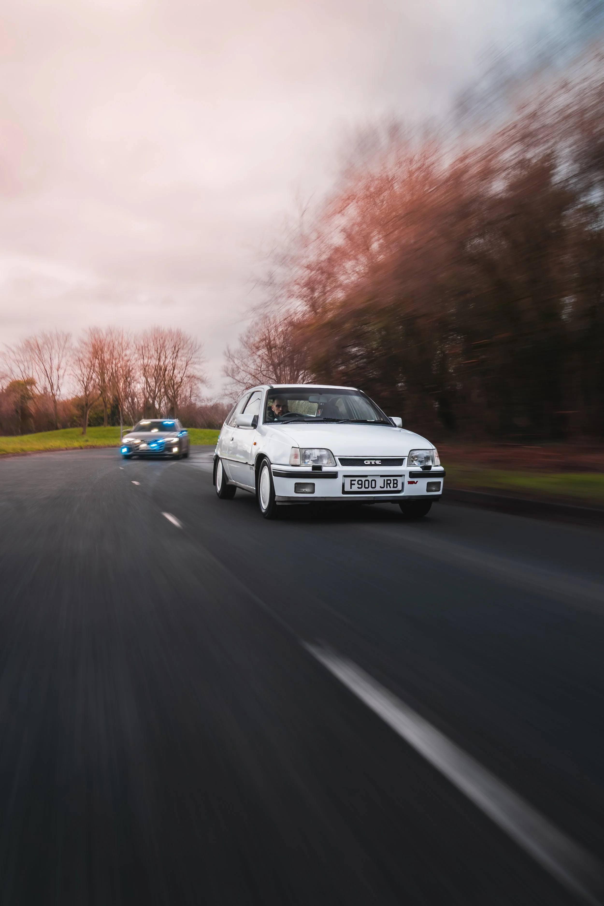 A white hatchback car with a UK license plate driving on a curved road, followed by a police car with flashing lights, with leafless trees and a cloudy sky in the background.