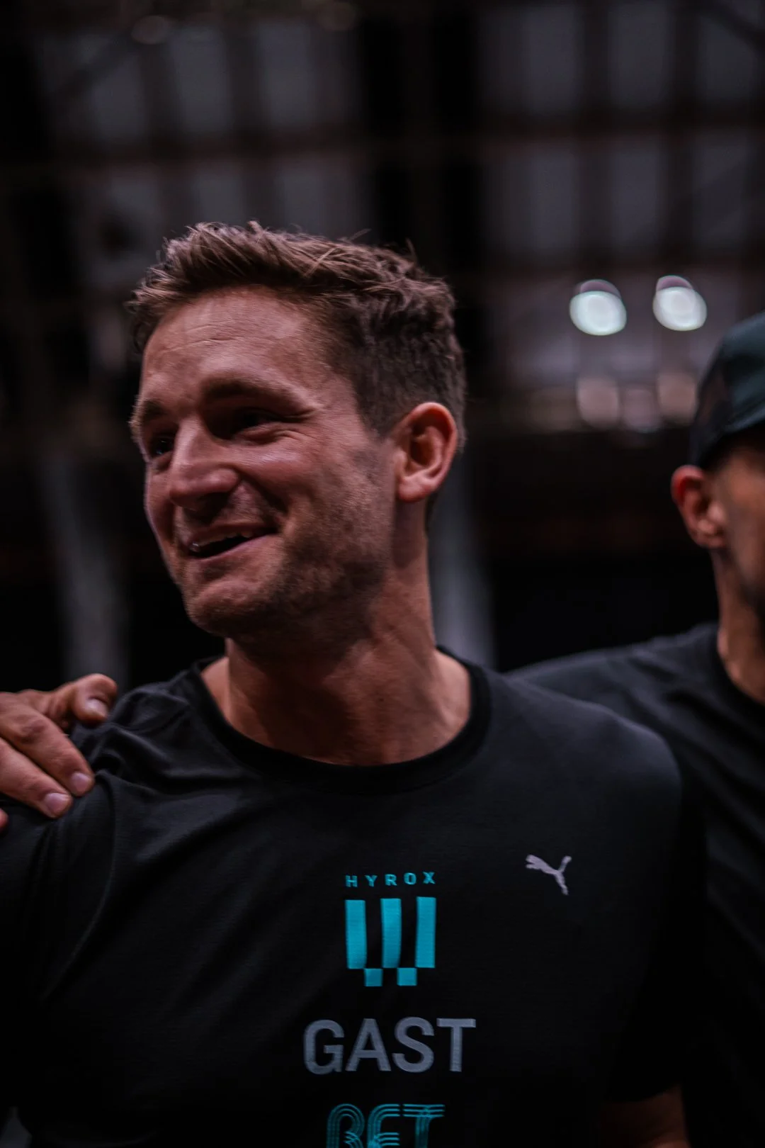 A man with short brown hair and a beard, smiling, wearing a black athletic shirt with logos, standing in a dark industrial or gym setting with another person partially visible beside him.