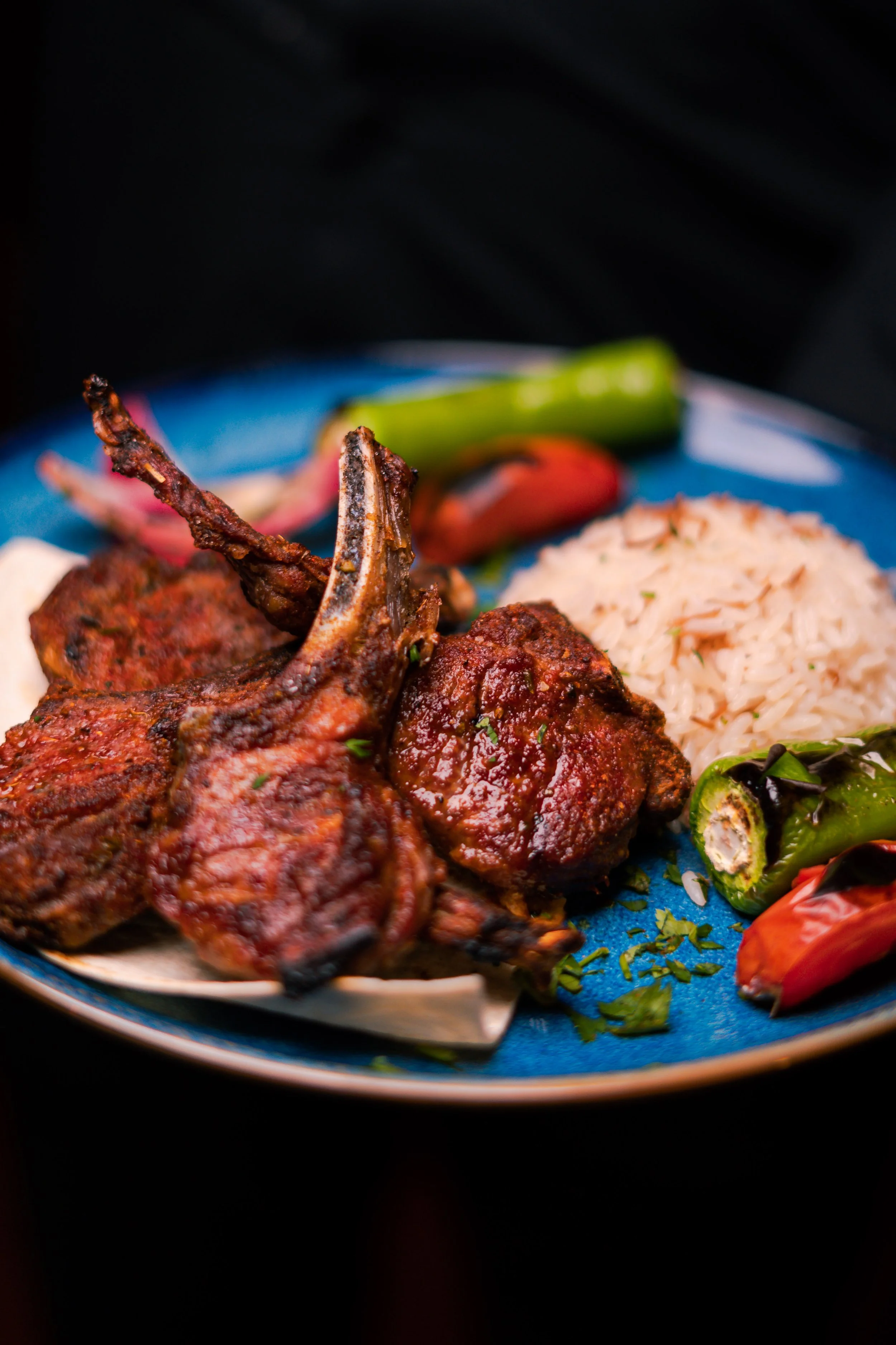 Close-up of grilled lamb chops, rice, and roasted peppers on a blue plate.