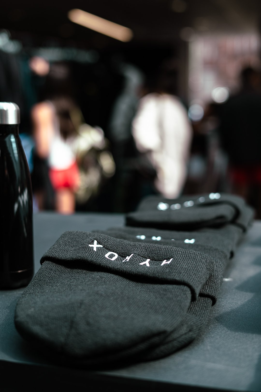 Stack of black athletic socks with white text on a table, blurred people in the background at an indoor event.