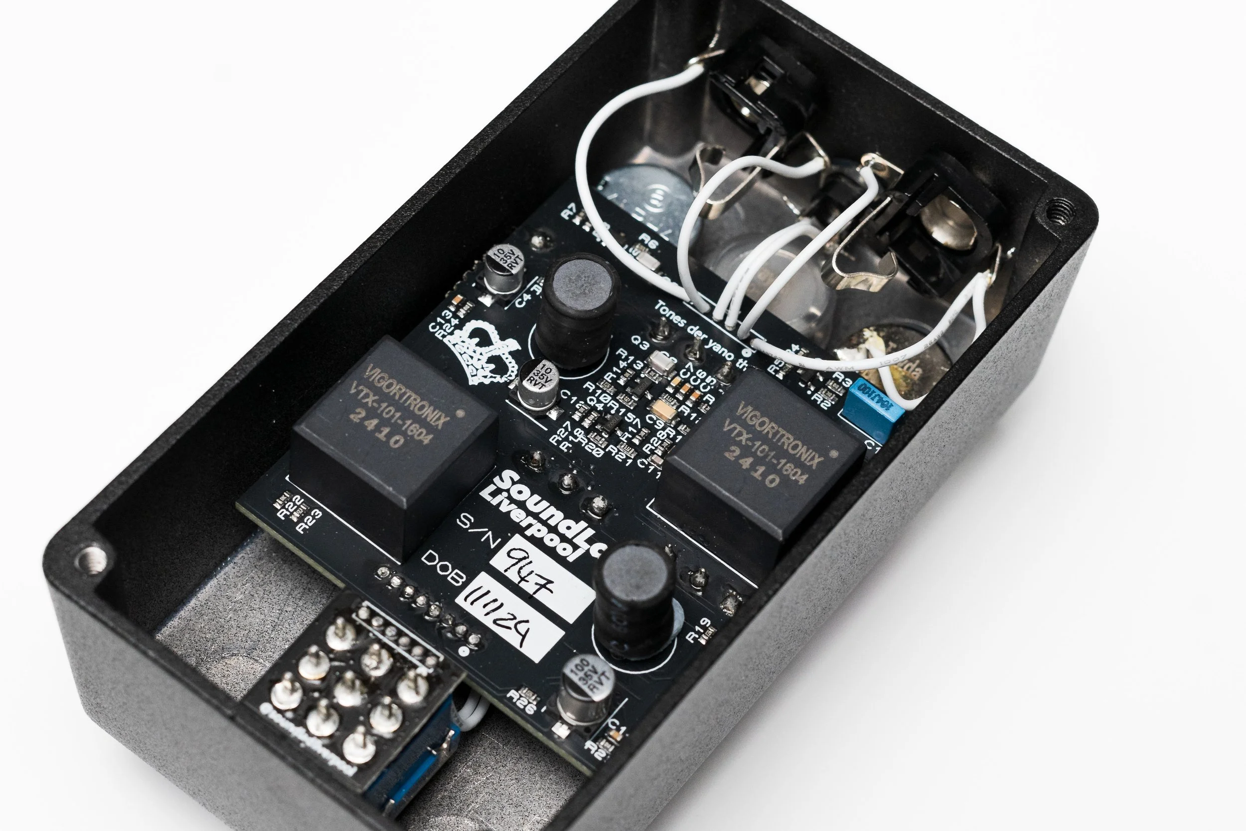 Inside view of an electronic guitar effects pedal showing circuit board, relays, capacitors, wires, and connectors.