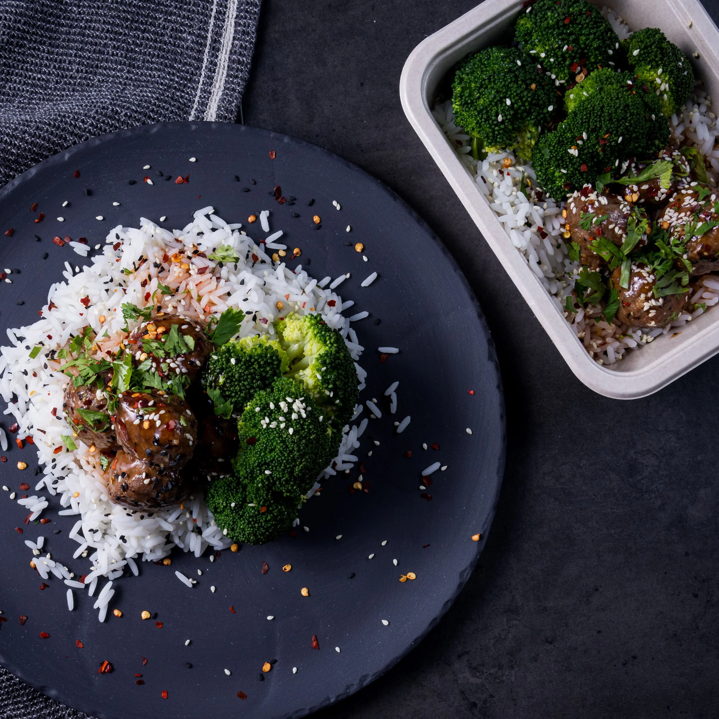 A black plate with white rice, broccoli, and meatballs topped with herbs and sesame seeds, with red pepper flakes and cilantro garnish. A container holds more broccoli and meatballs on a dark surface.