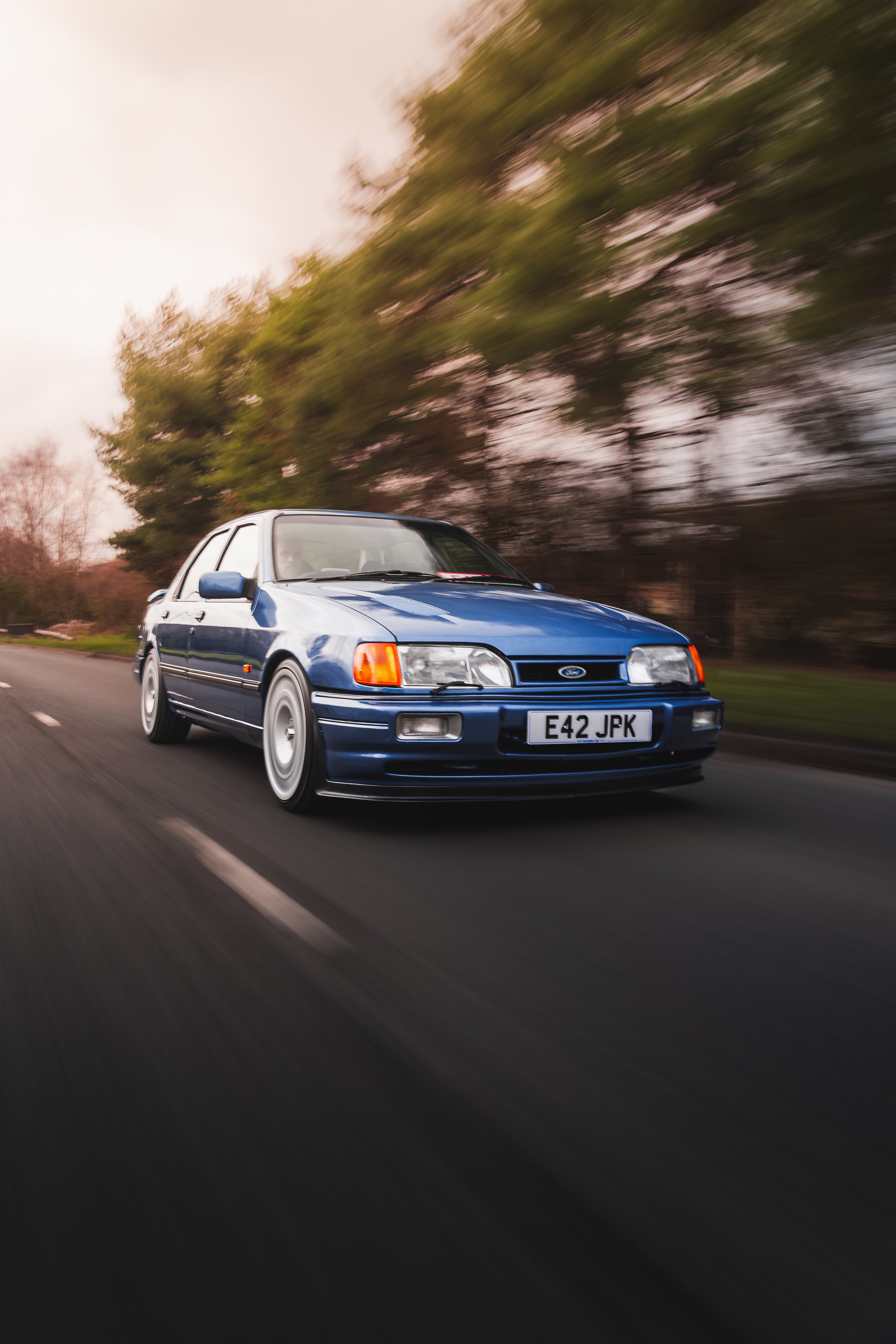 A blue vintage Ford car driving on a road with blurred trees in the background