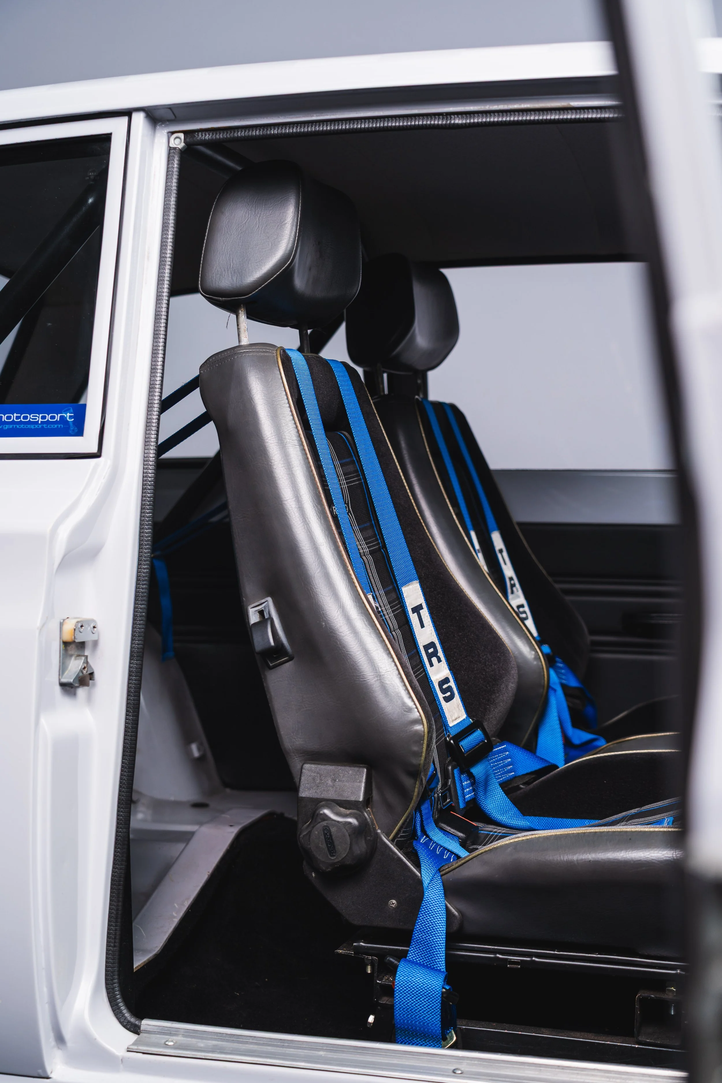 The interior of a racing vehicle showing two black leather seats with blue harnesses marked 'RS'.