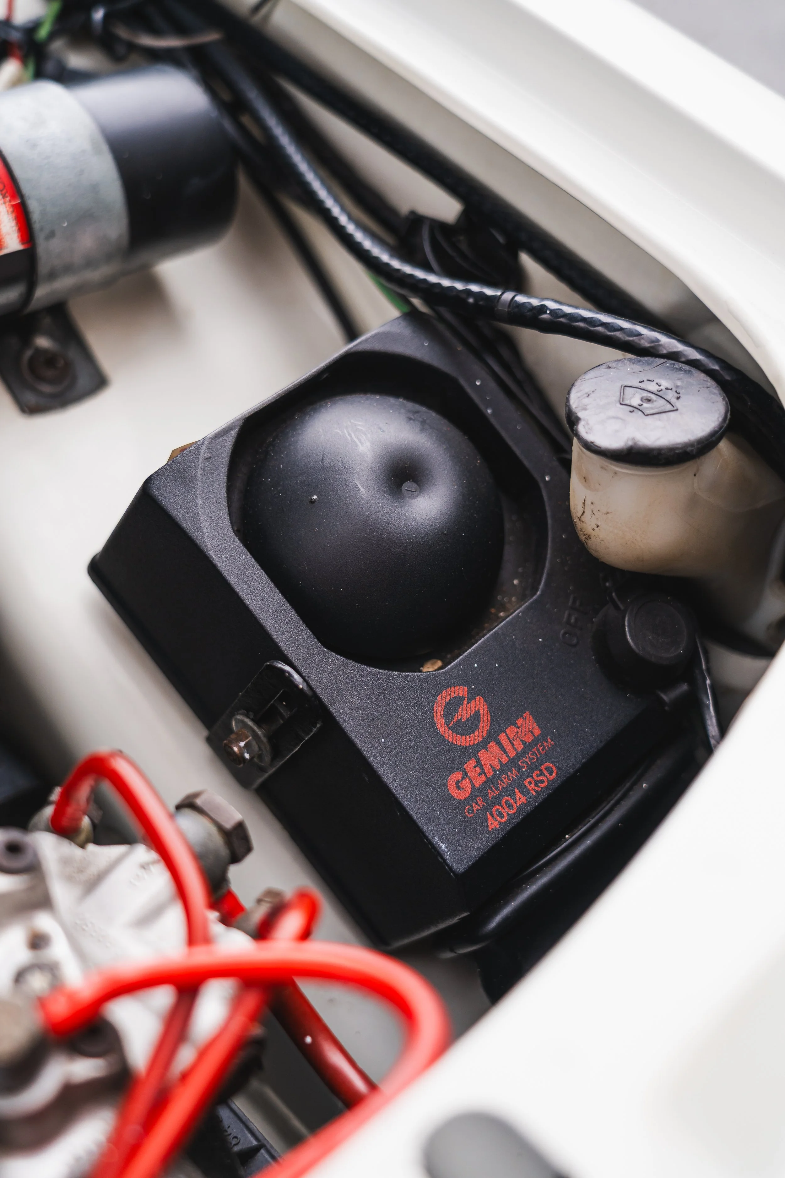 Close-up of a Gemlux car alarm system mounted in a vehicle's engine compartment, with various wiring and other engine components visible.