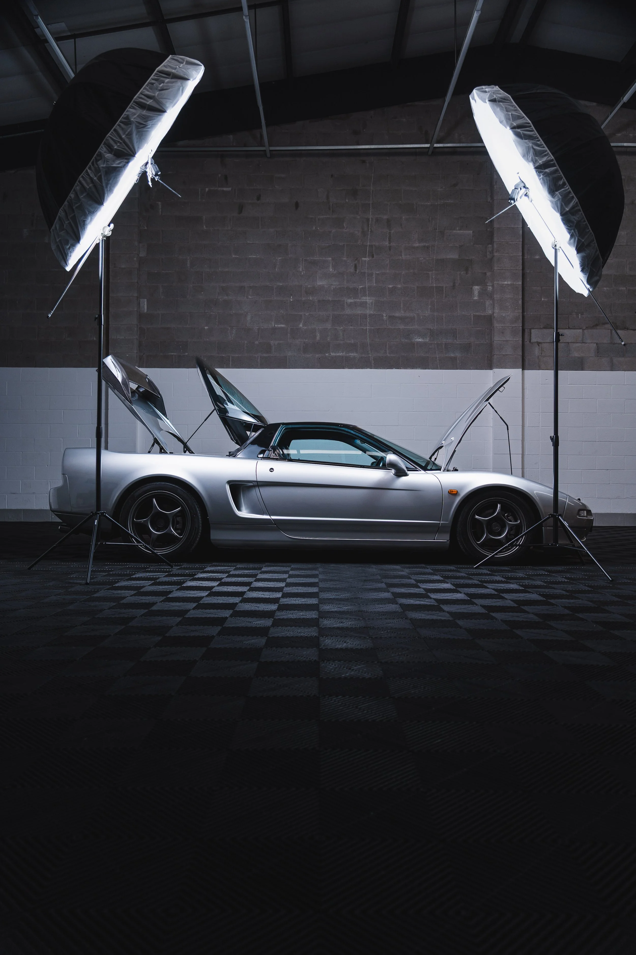 Silver sports car with open doors and hood, set up in a photography studio with two large softbox lights overhead.