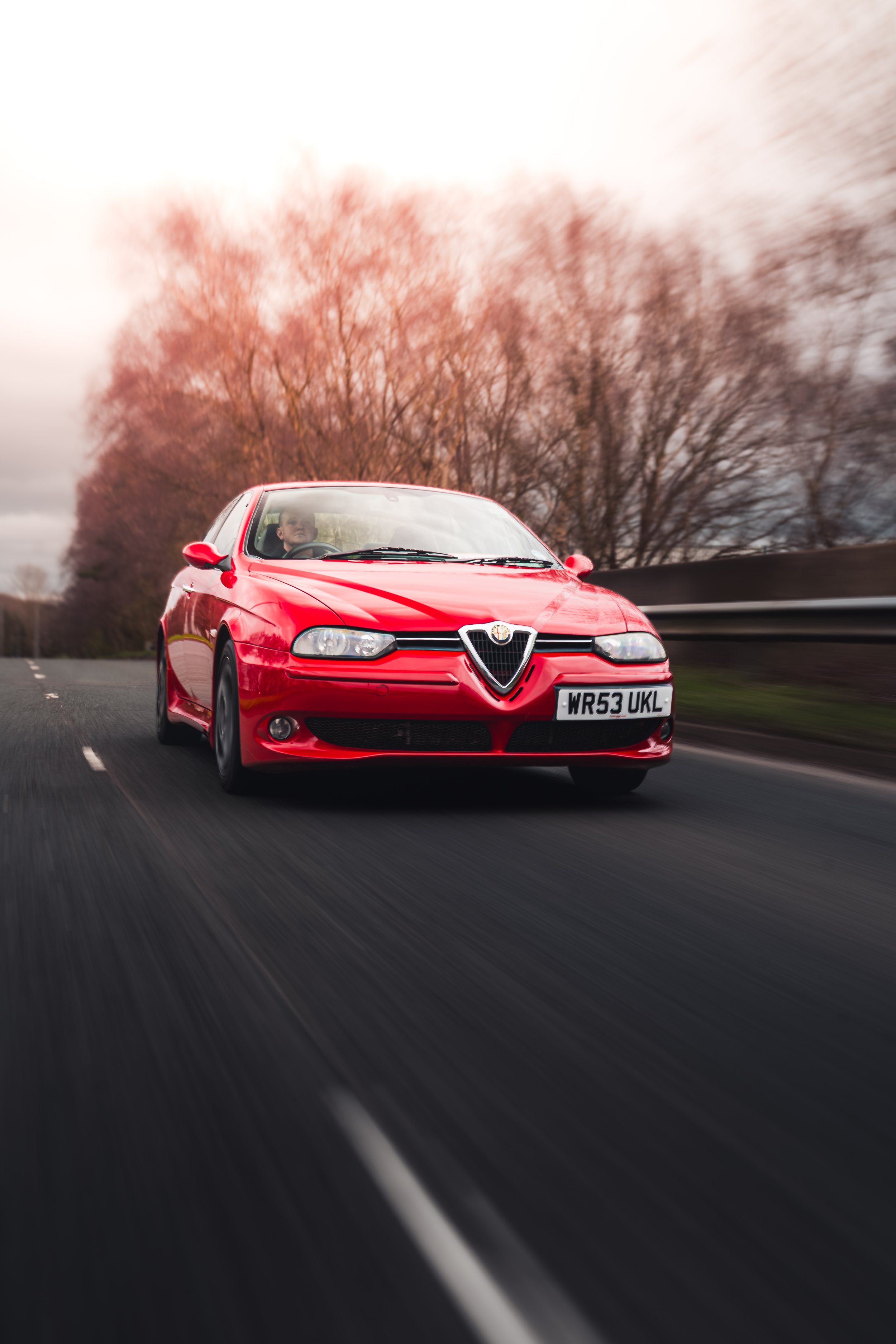 Red Alfa Romeo car driving on a highway with pink trees and cloudy sky in the background.