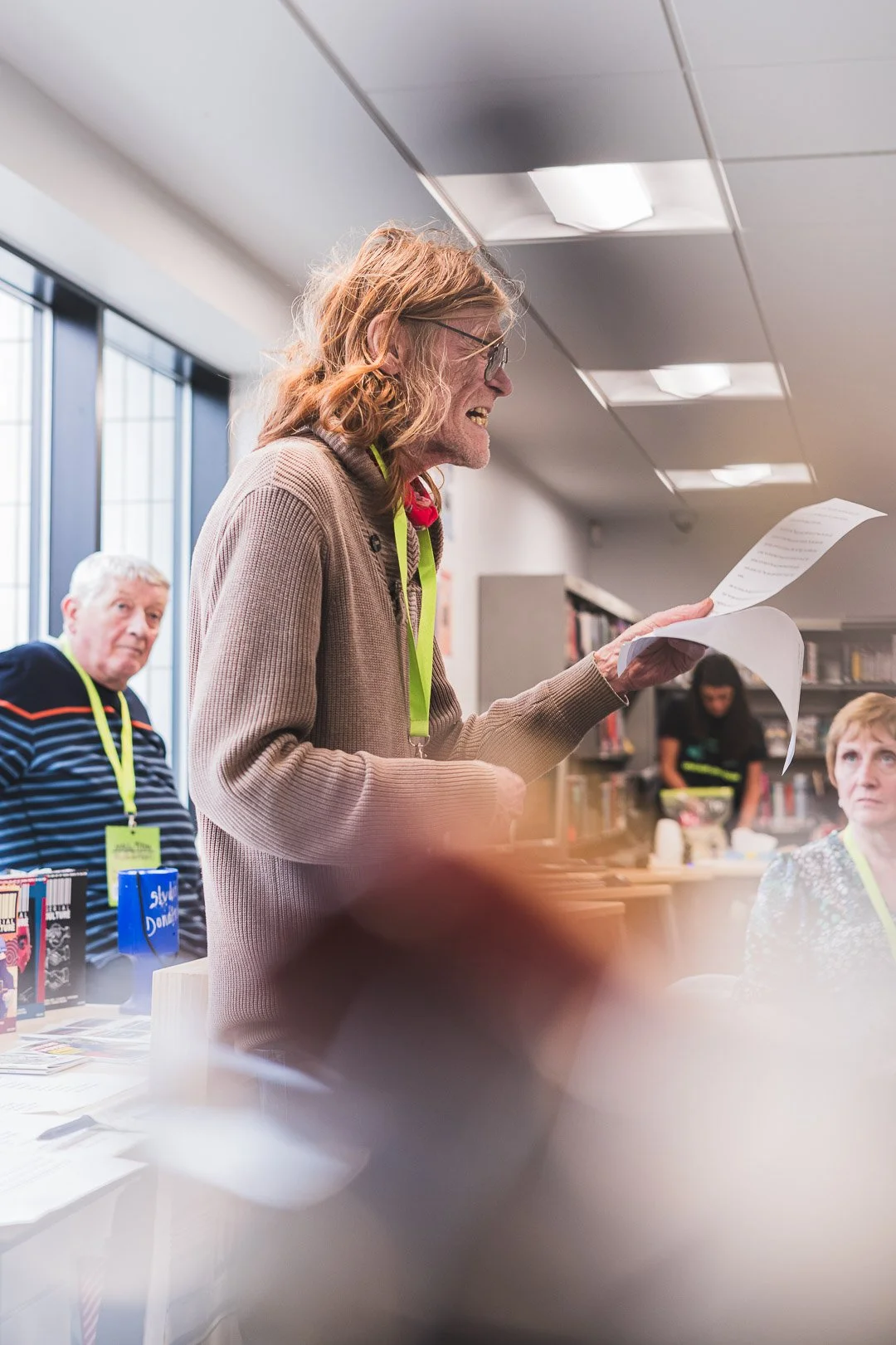 A woman in glasses and a beige sweater stands in a library, reading from a paper, with people in the background.