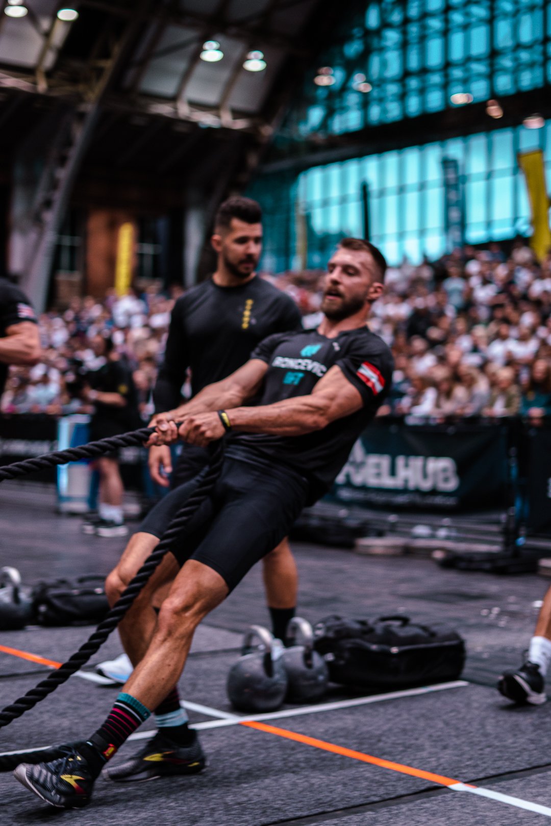 A man in black athletic clothing and colorful socks participates in a tug-of-war competition at a fitness event, with others and a large audience watching in the background.