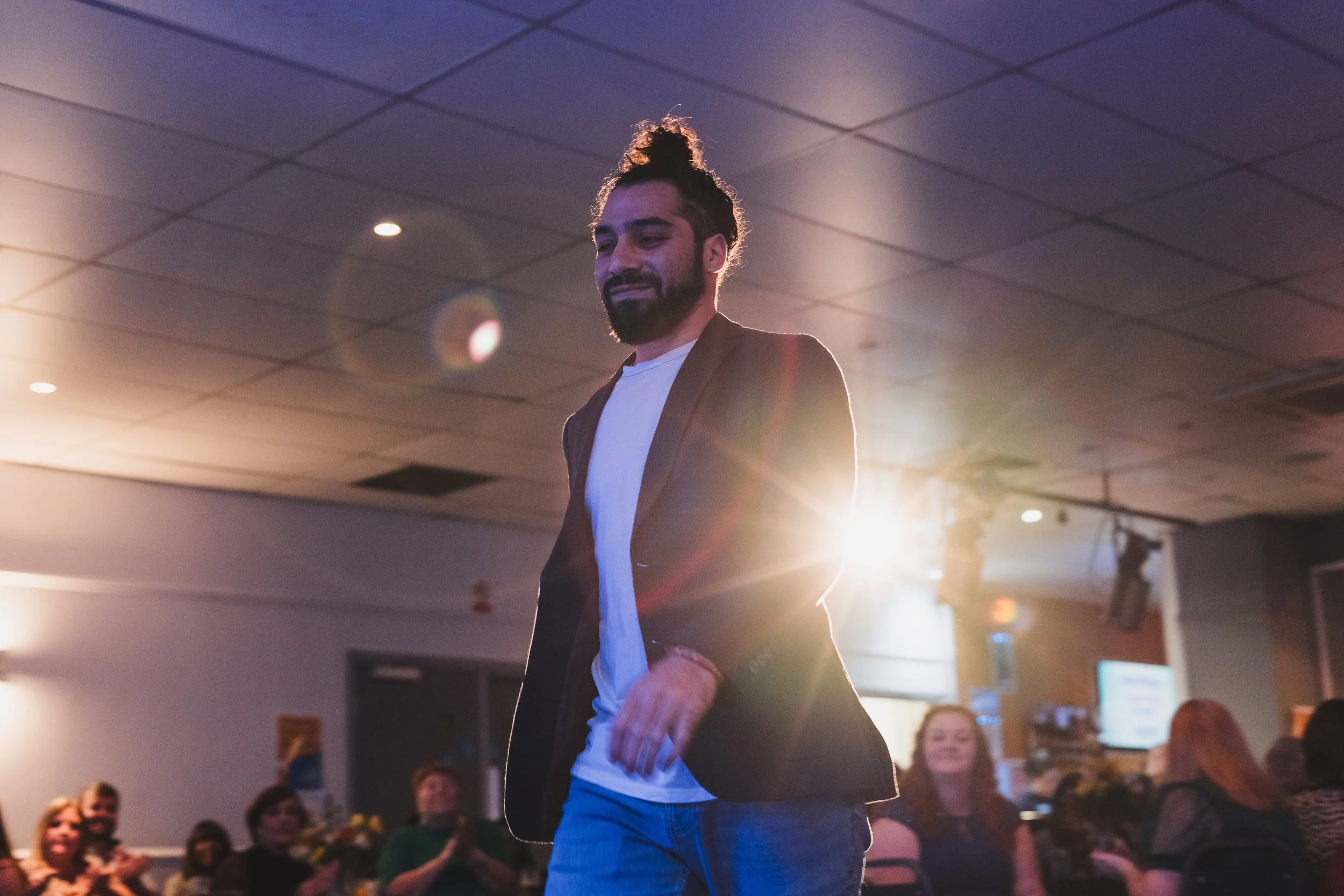 A man with a beard and curly hair styled in a top knot walking in a dimly lit room with an audience watching, illuminated by bright backlight.