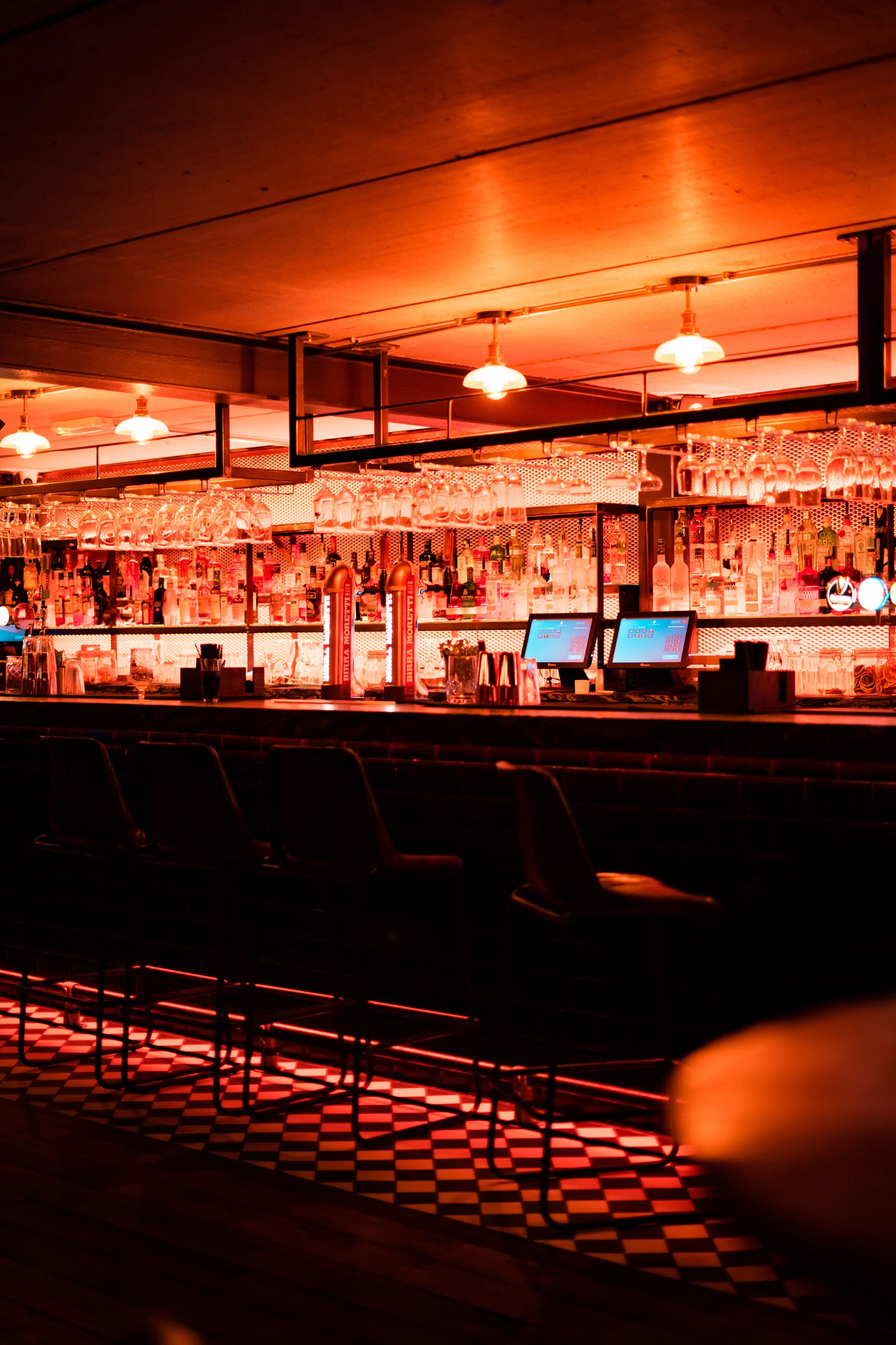 A dimly lit bar with a glowing pink hue, bar stools, and a counter with bottles and glassware.
