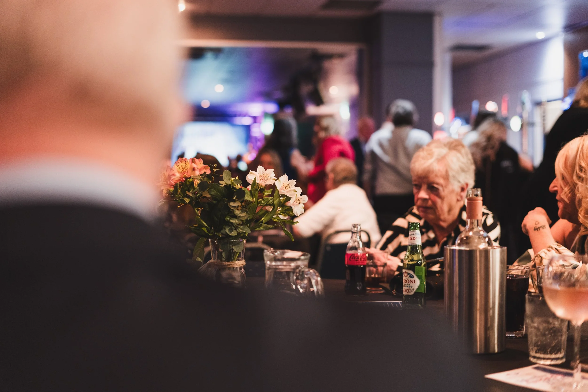 People gathered at a social event or restaurant with drinks, flowers, and a dark interior with colorful lights, some are sitting at tables, and there is a busy background.
