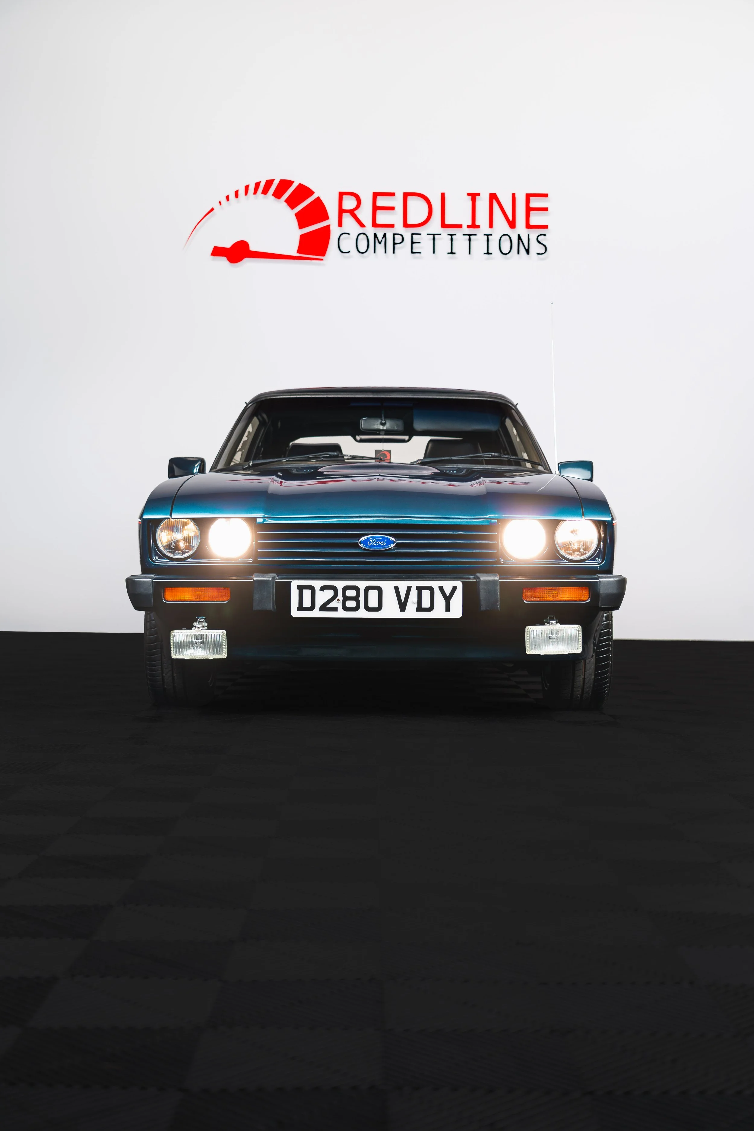 Front view of a classic black Ford car with headlights on, parked in a setting with a white background. Above the car is the logo for 'Redline Competitions' featuring a speedometer graphic.
