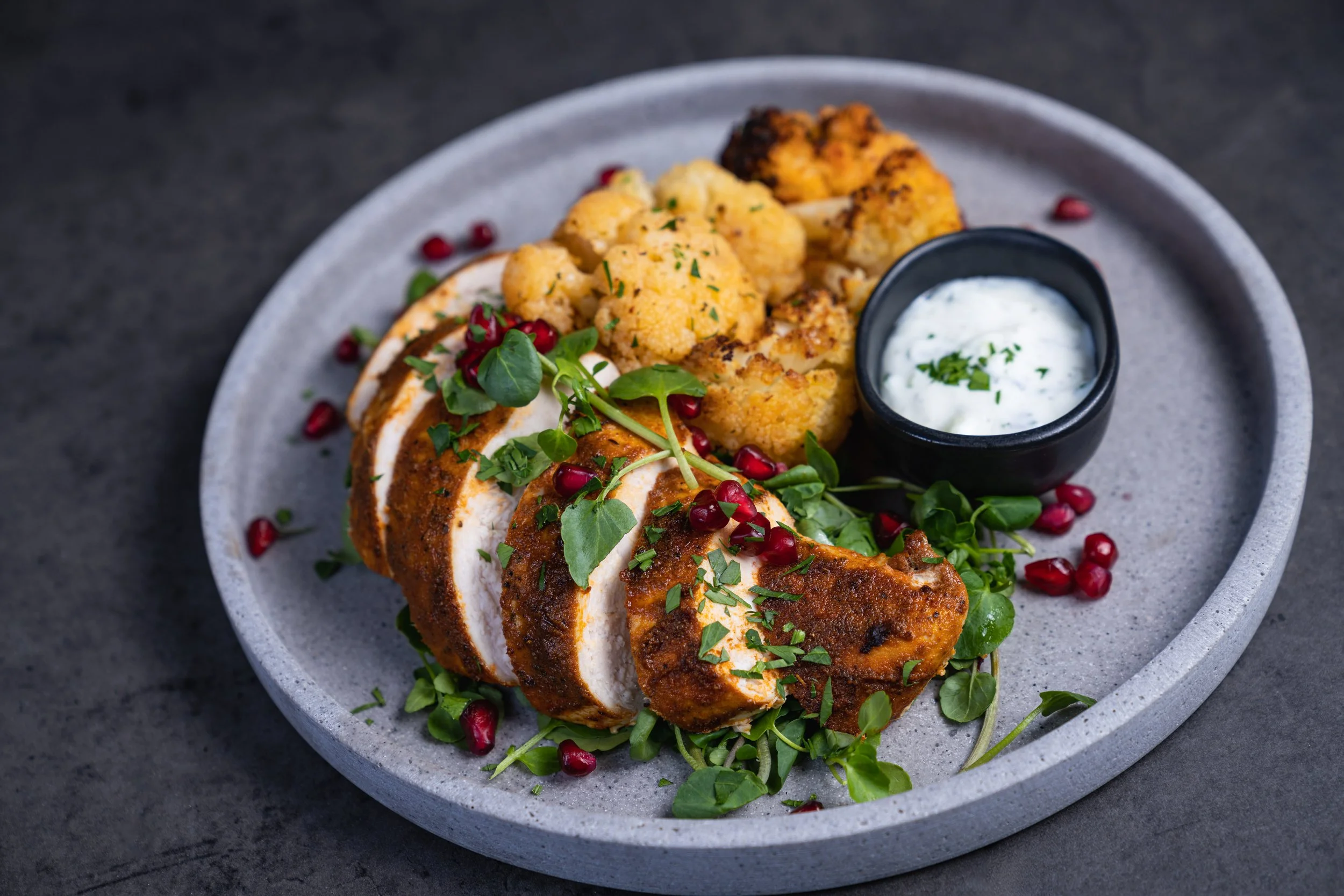 Plate with sliced roasted chicken, roasted cauliflower, pomegranate seeds, microgreens, and a small bowl of creamy sauce garnished with herbs on a gray plate.