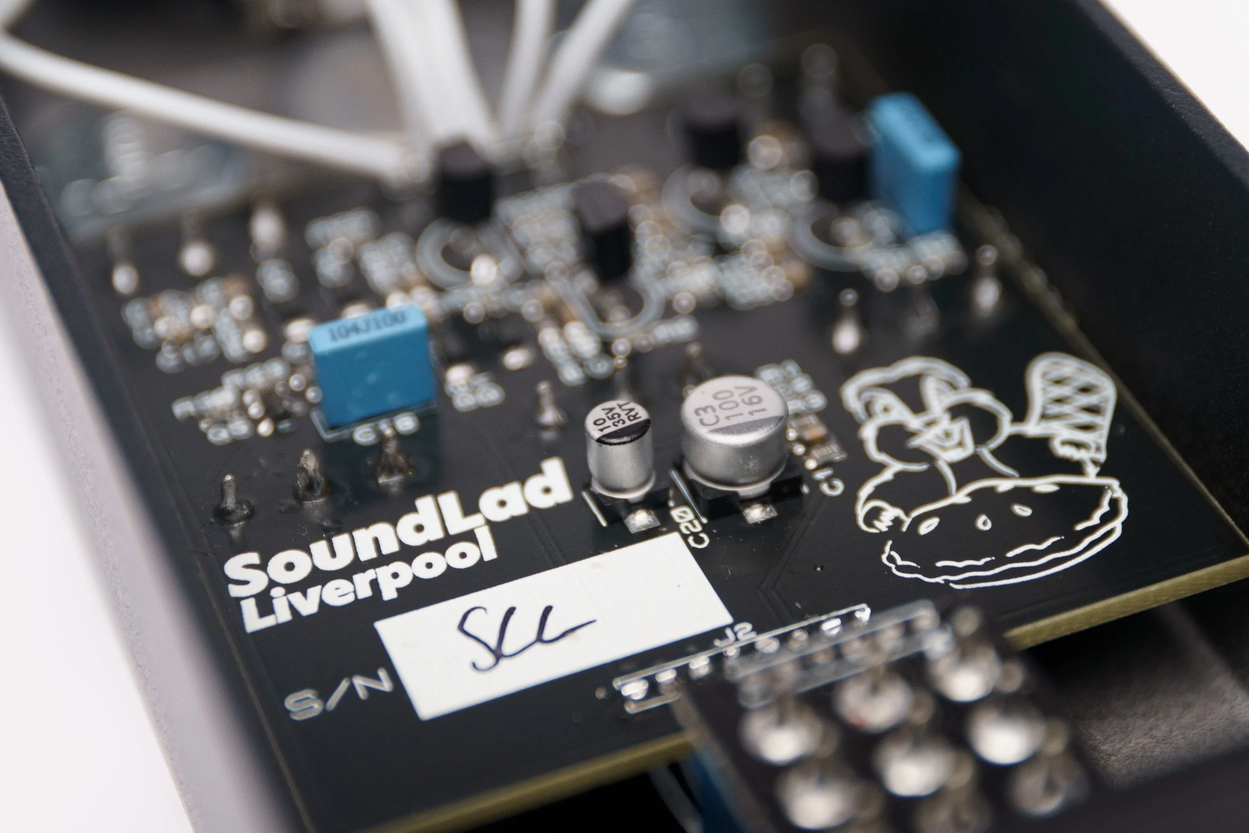 Close-up of a black circuit board labeled 'SoundLab Liverpool' with electronic components, including capacitors, connectors, and a White cartoon character holding a pizza drawing.