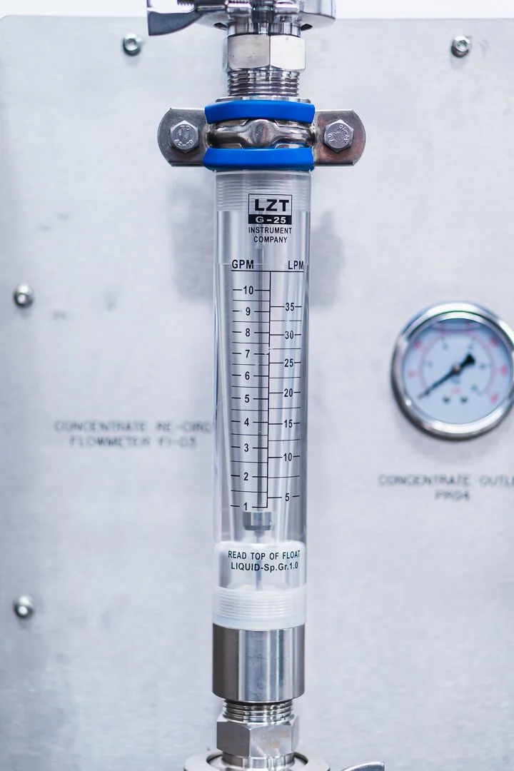 A clear liquid flow meter with blue clamps and metal fittings, mounted on a metal surface, with pressure gauge on the right side of the image.