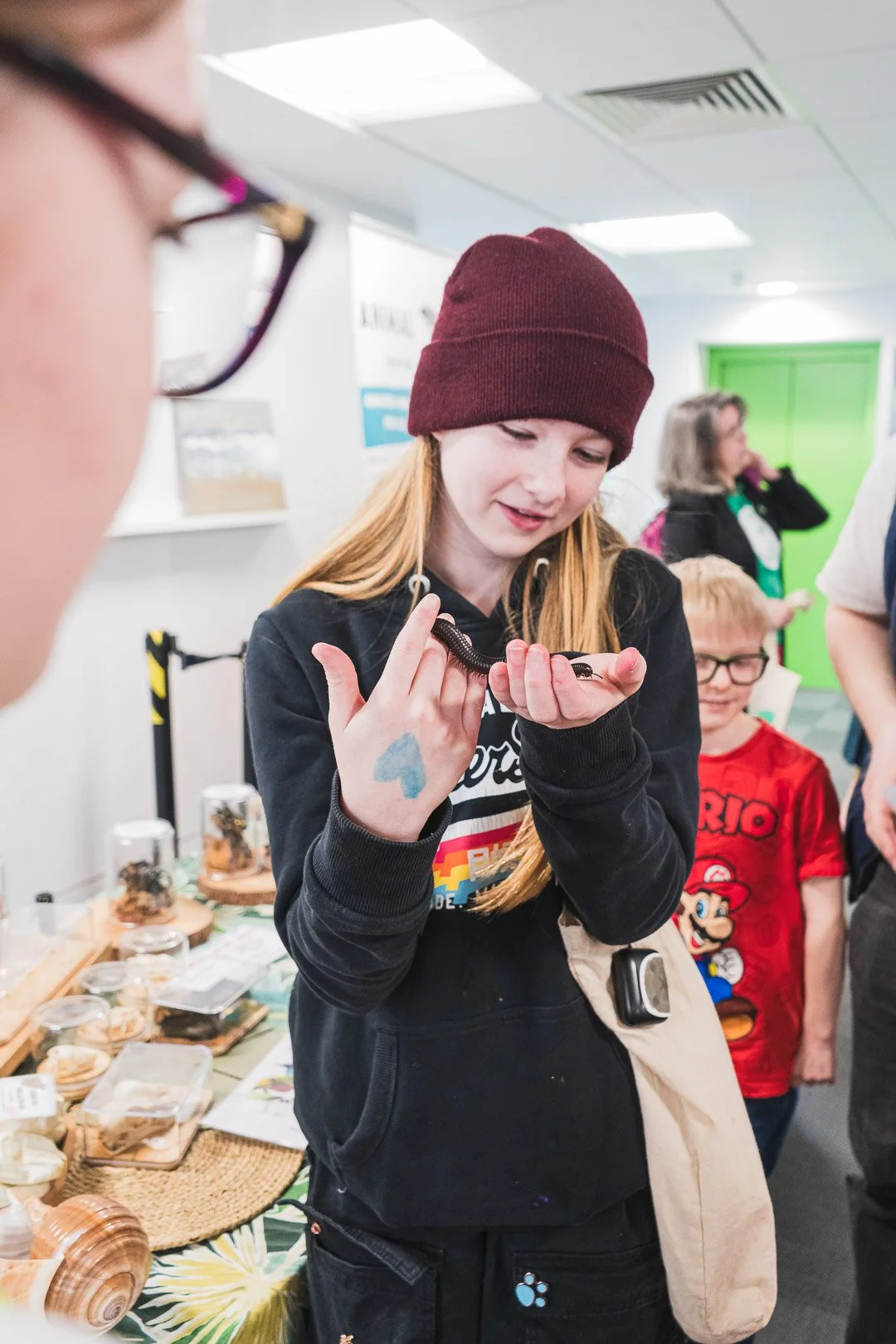 A young girl with long red hair, wearing a maroon beanie and black hoodie, is holding a large black centipede and examining it closely. Other children and adults are in the background at an indoor event with display tables.