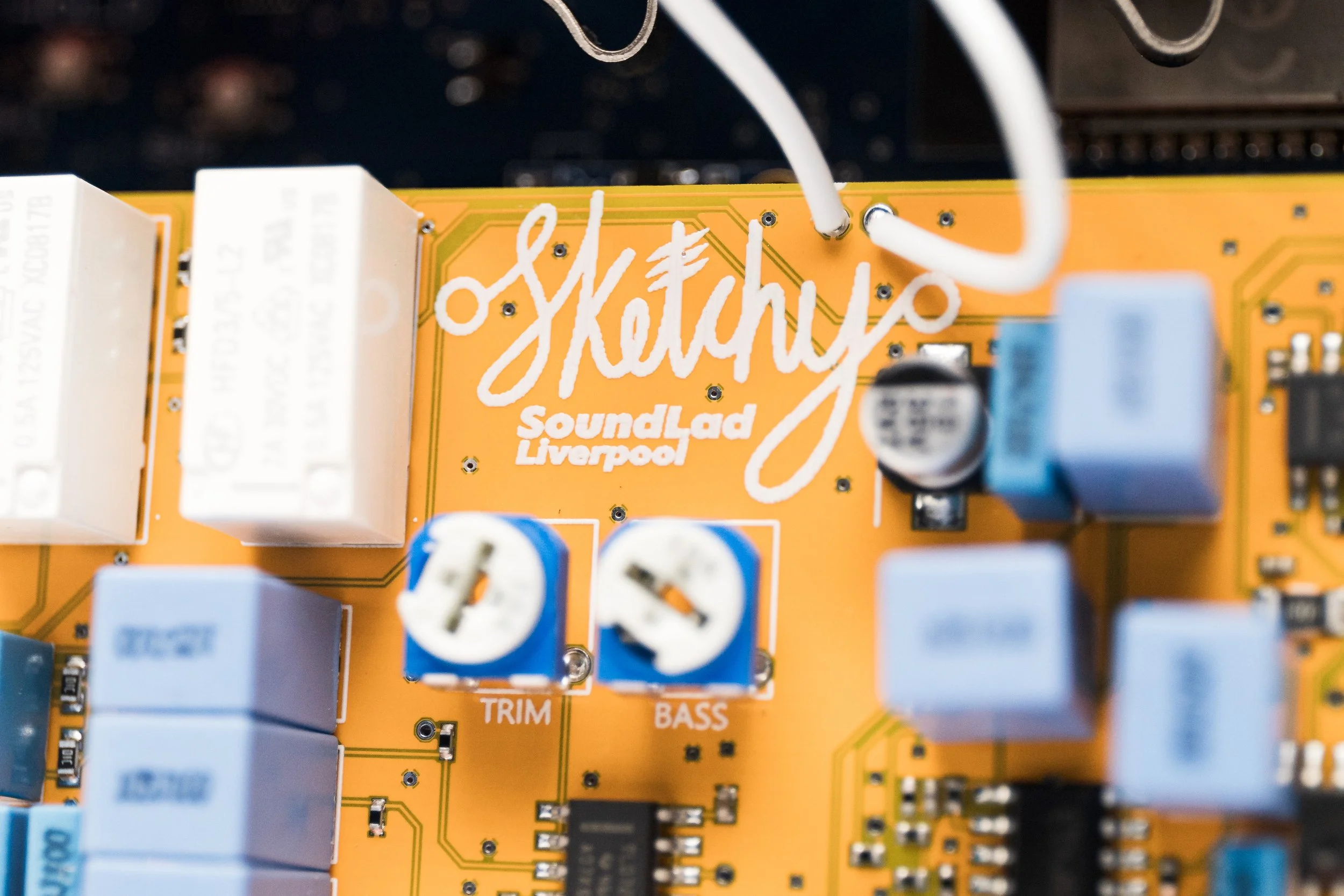 Close-up of a yellow circuit board with the word "Sketchy" in white cursive and "SoundLad Liverpool" in smaller text below. Various electronic components, including blue and white knobs labeled "Trim" and "Bass," are attached to the board.