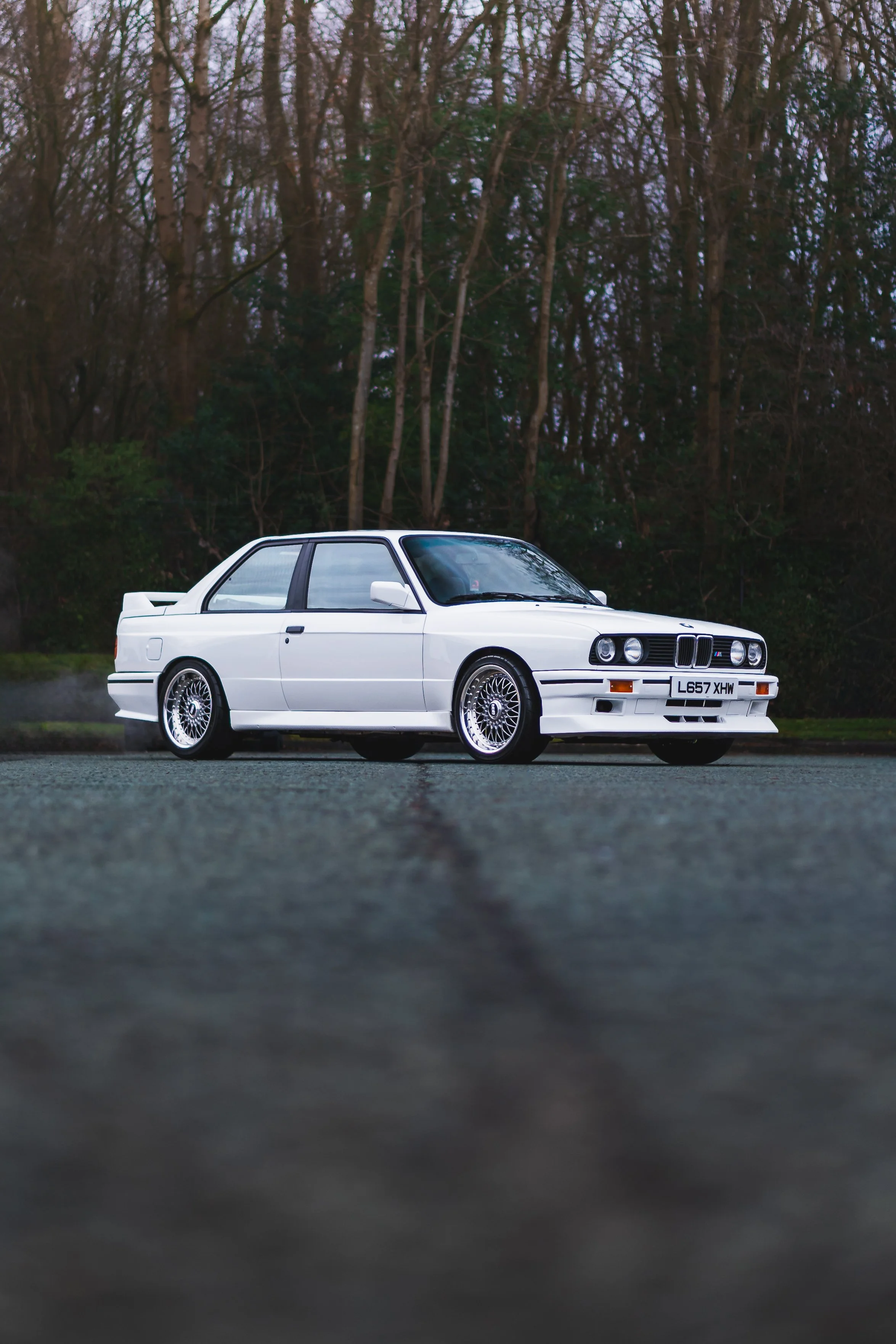 A white vintage BMW E30 M3 parked outdoors on a paved surface with a background of leafless trees, during the evening or early morning.