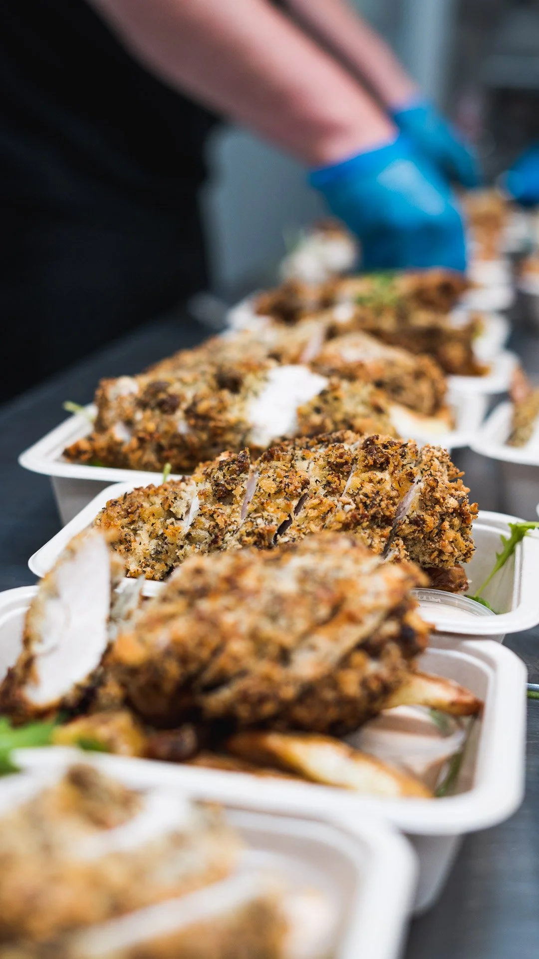 Multiple breaded and fried chicken pieces in white takeout containers on a black surface, with a person in the background wearing blue gloves preparing or serving food.