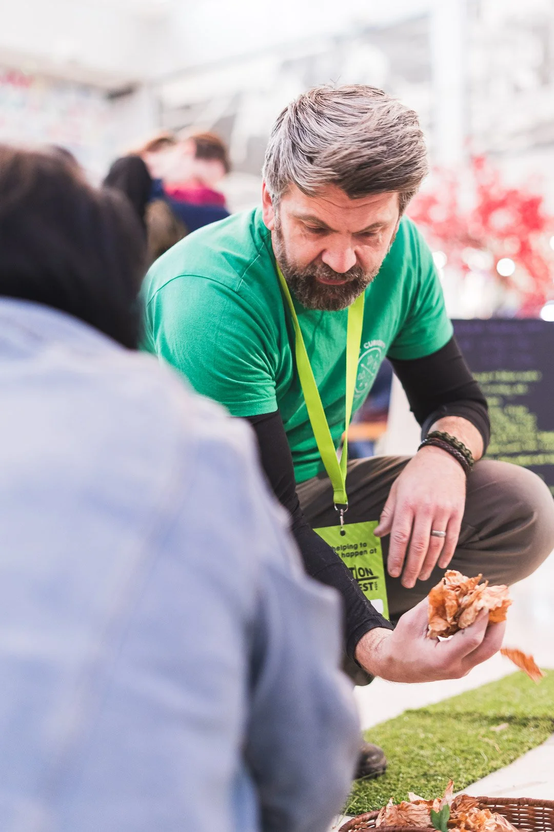 A man with gray hair and a beard wearing a green t-shirt kneels down and shows a piece of apple peel to a person with dark hair, in an indoor setting with several people in the background.