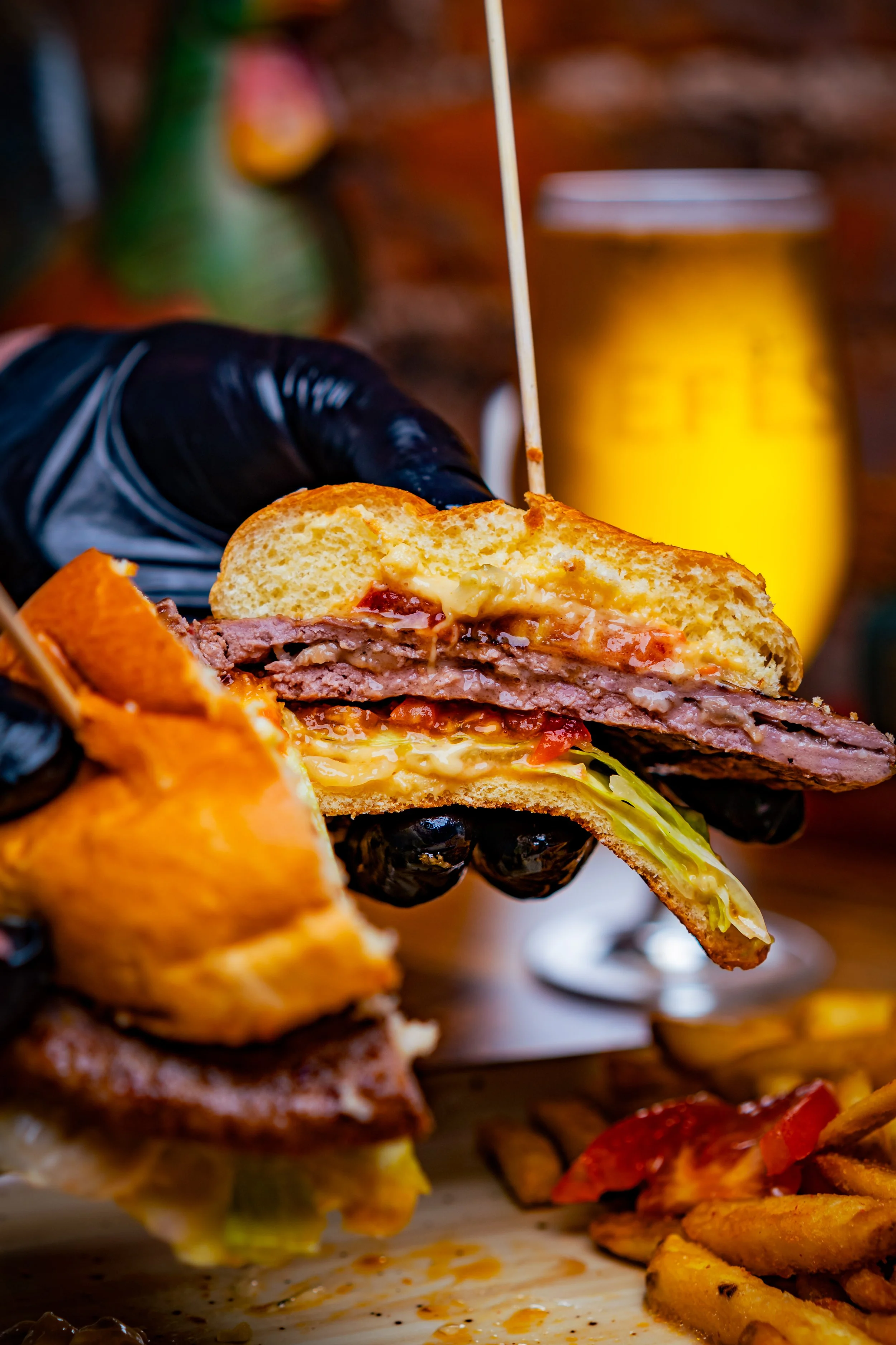Close-up of a partially eaten double cheeseburger with lettuce, tomato, cheese, and beef patty, held by a person wearing a black glove, with a glass of beer and fries in the background.