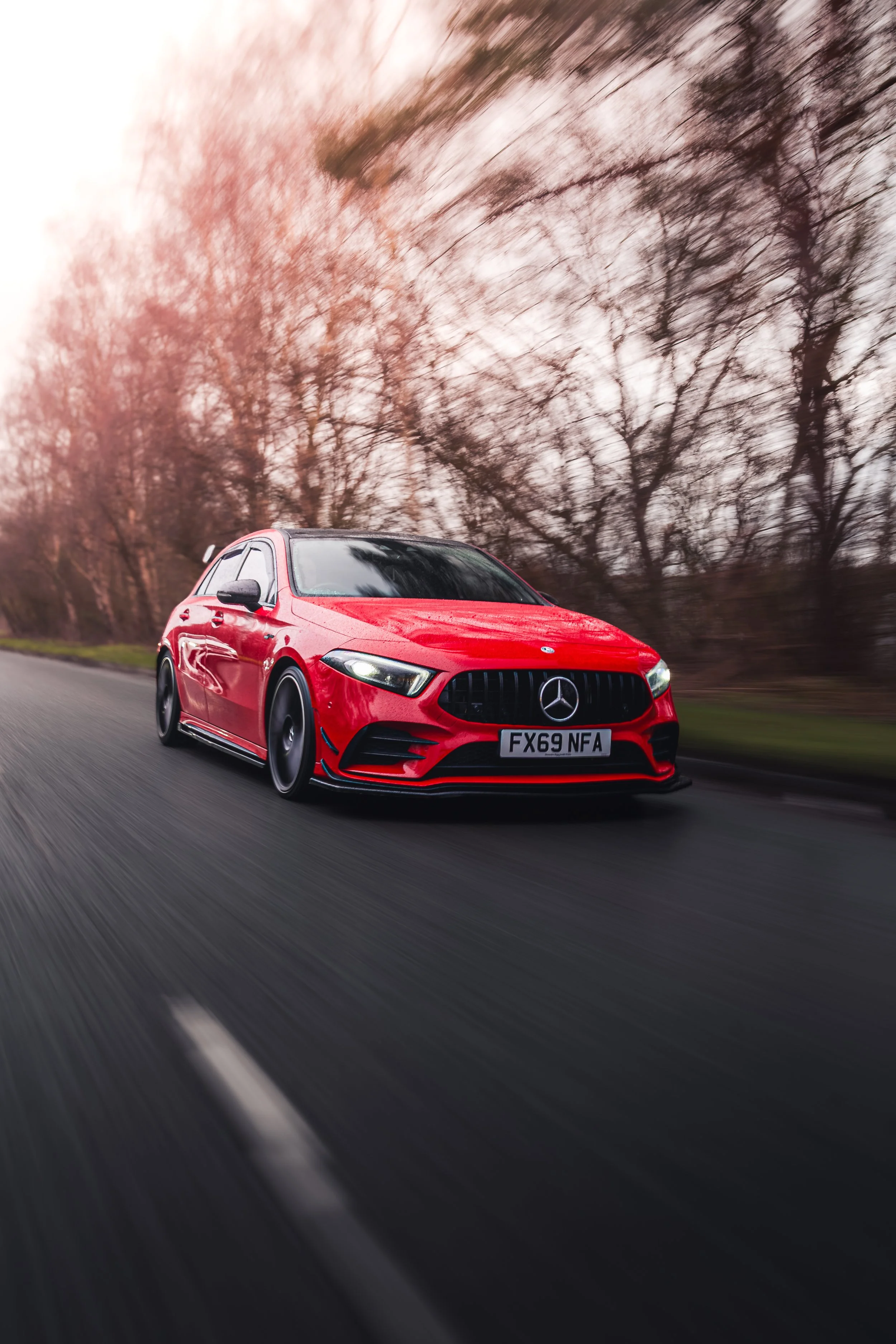 A red Mercedes-Benz car driving on a road with a blurred background of leafless trees and a pinkish sky.