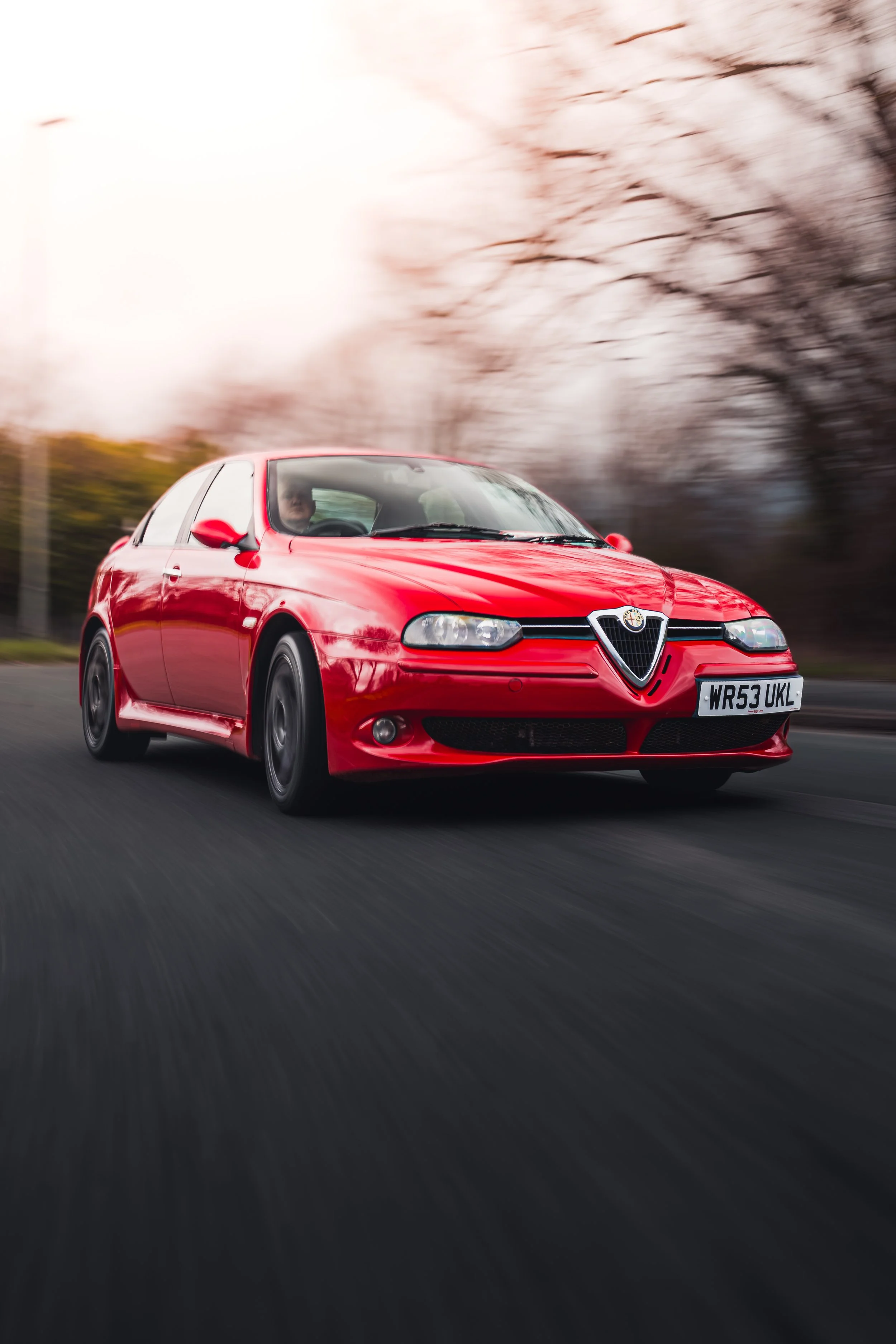 Red Alfa Romeo sedan driving on a road with blurred trees in the background.